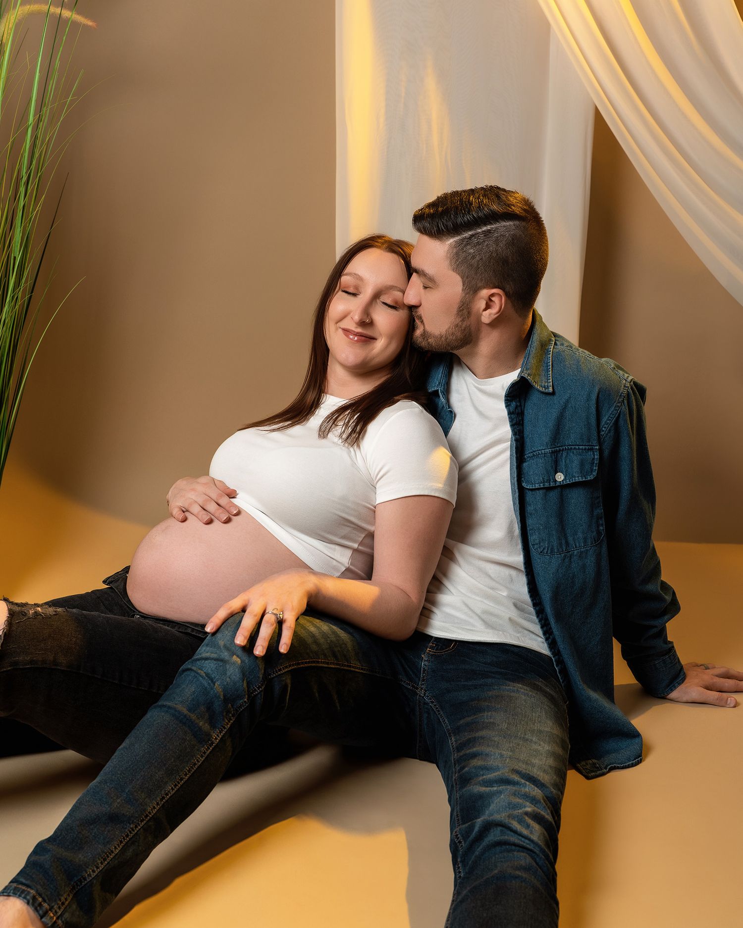 Casual maternity portrait with couple in denim and white clothing on yellow backdrop.