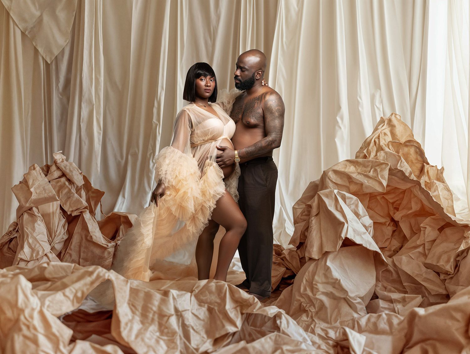 Artistic maternity photo with couple posing against beige fabric backdrop with crumpled paper elements.