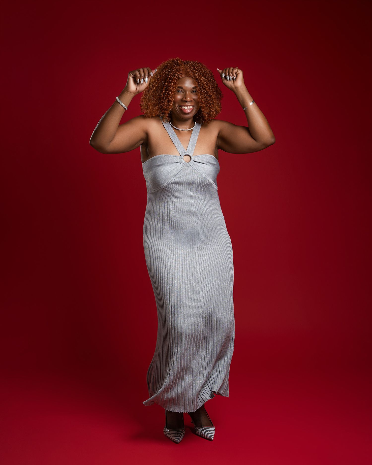 Joyful pose in silver dress against red backdrop with dramatic lighting.