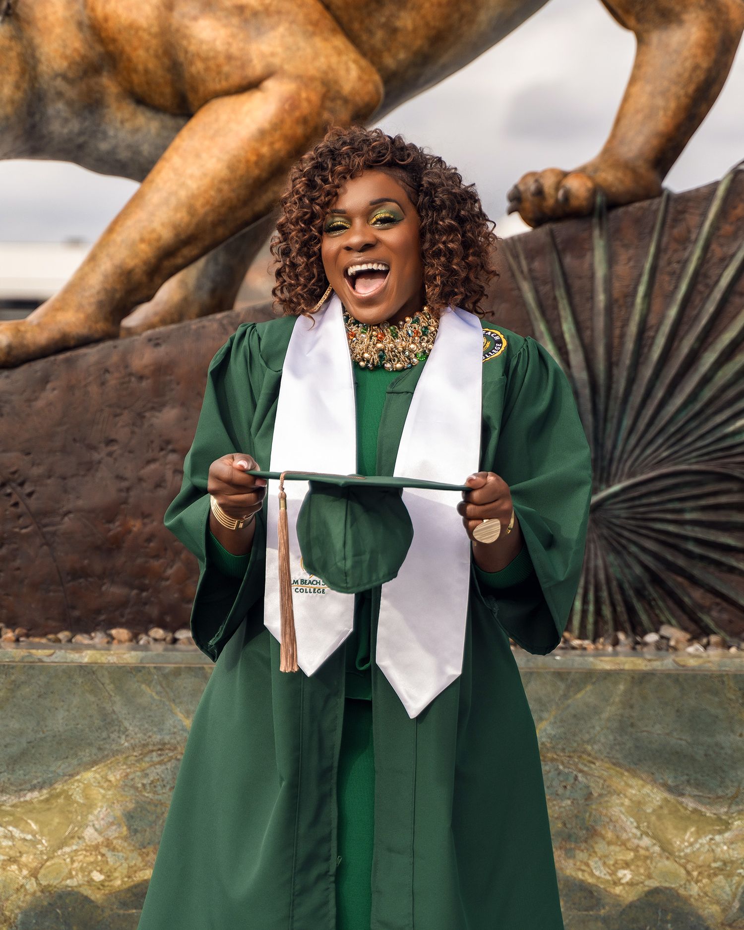 Graduate holding diploma poses joyfully beneath bronze wildcat statue.