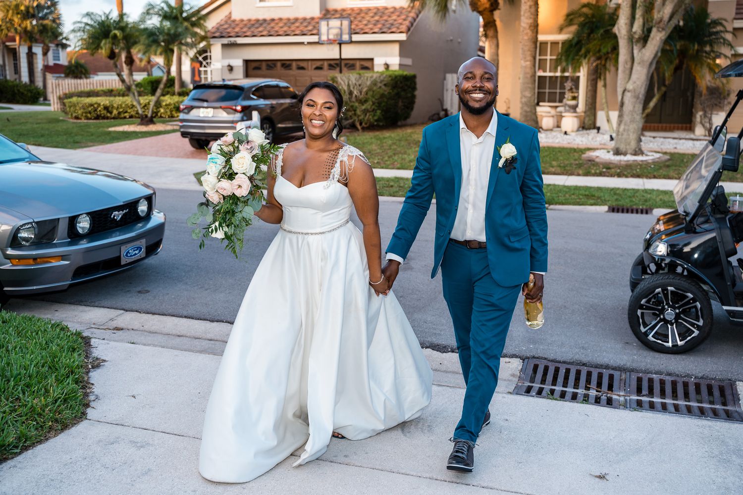 Newlyweds walk together outdoors, bride in white gown and groom in blue suit carrying a floral bouquet.
