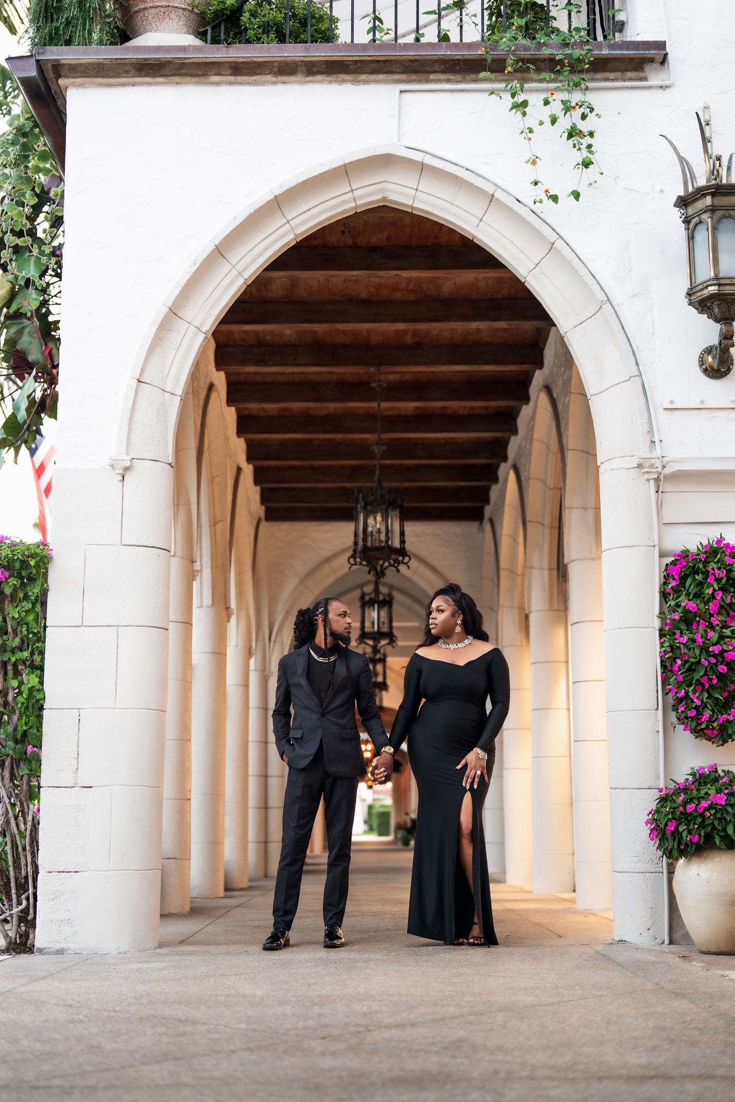 A stylish couple stands together under a grand arched hallway adorned with flowering vines and lanterns.