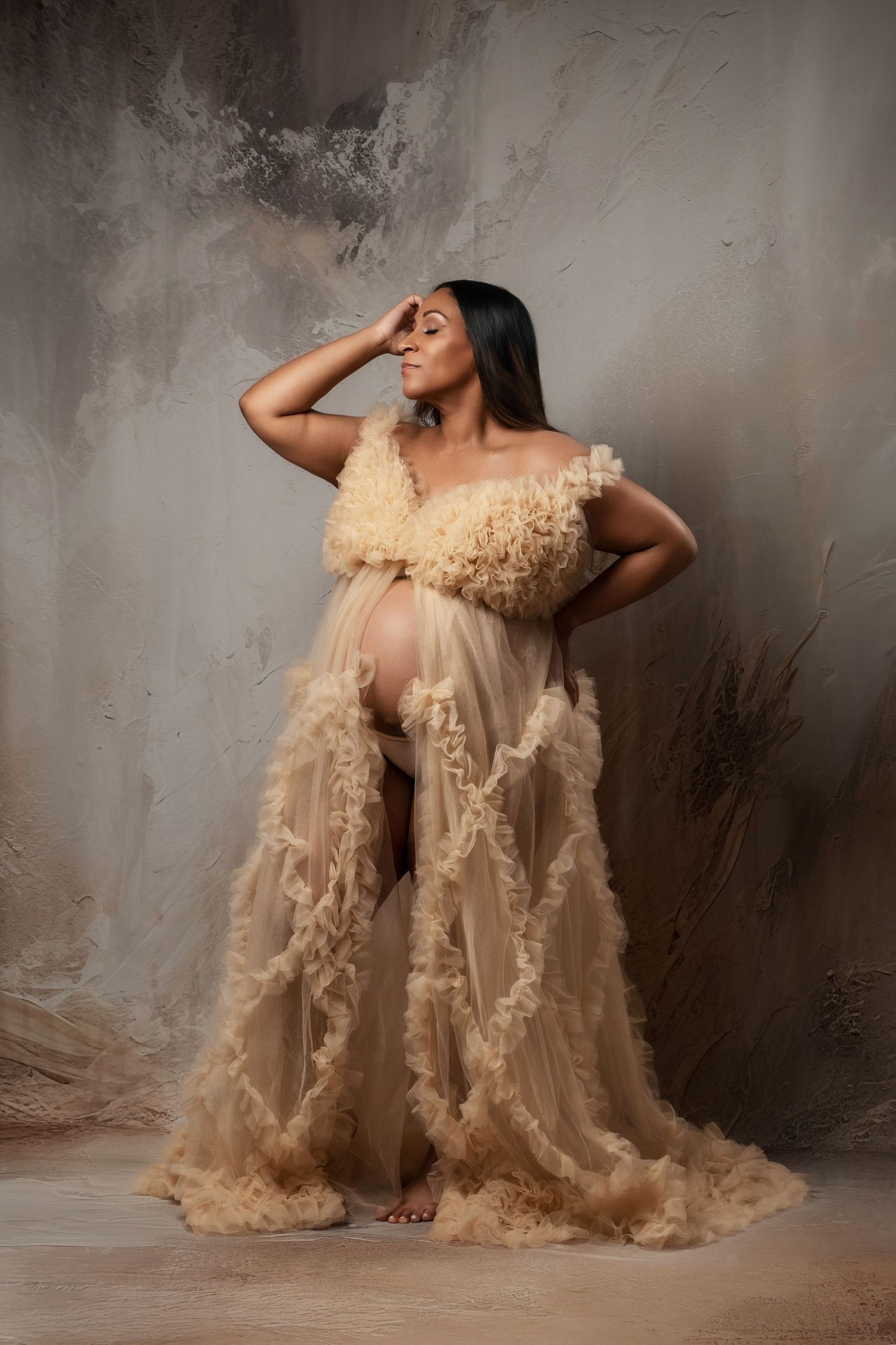 Artistic maternity photoshoot featuring flowing champagne-colored tulle gown against textured grey backdrop.