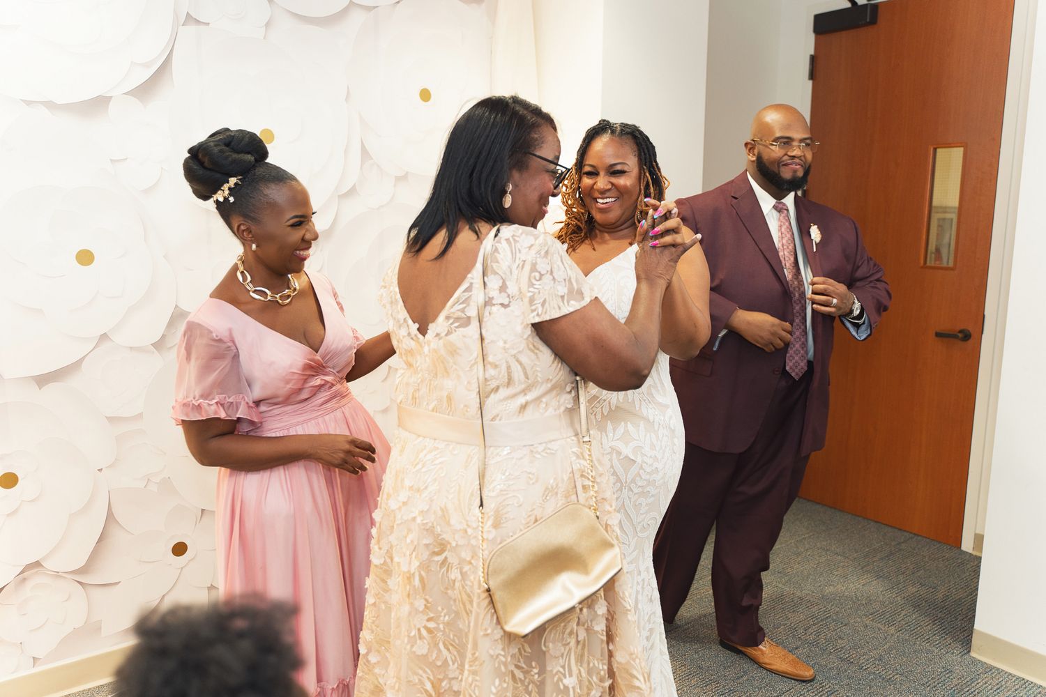 Wedding guests sharing emotional embraces and laughter in a hallway with decorative white wall art.