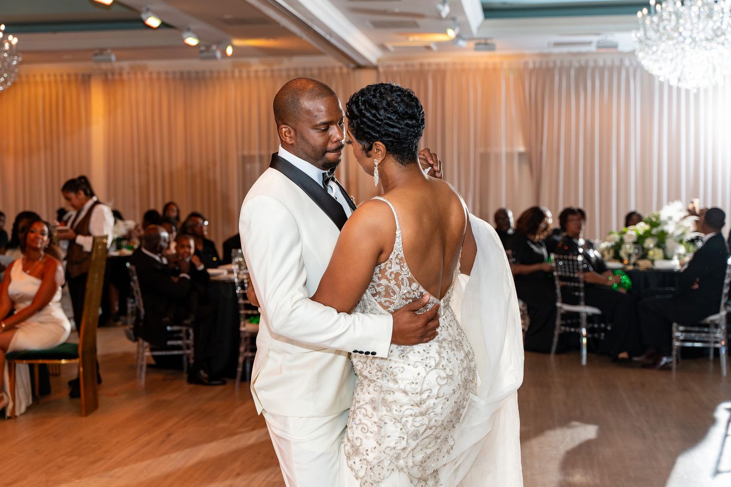 Couple shares an intimate slow dance moment in an elegant reception hall with crystal chandeliers.
