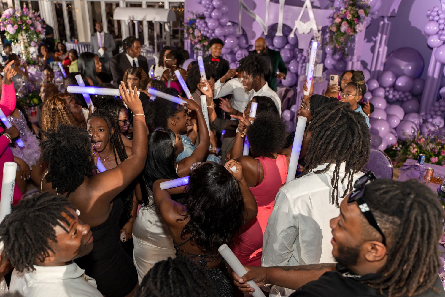 Party guests dancing and celebrating under purple lighting with balloon decorations.