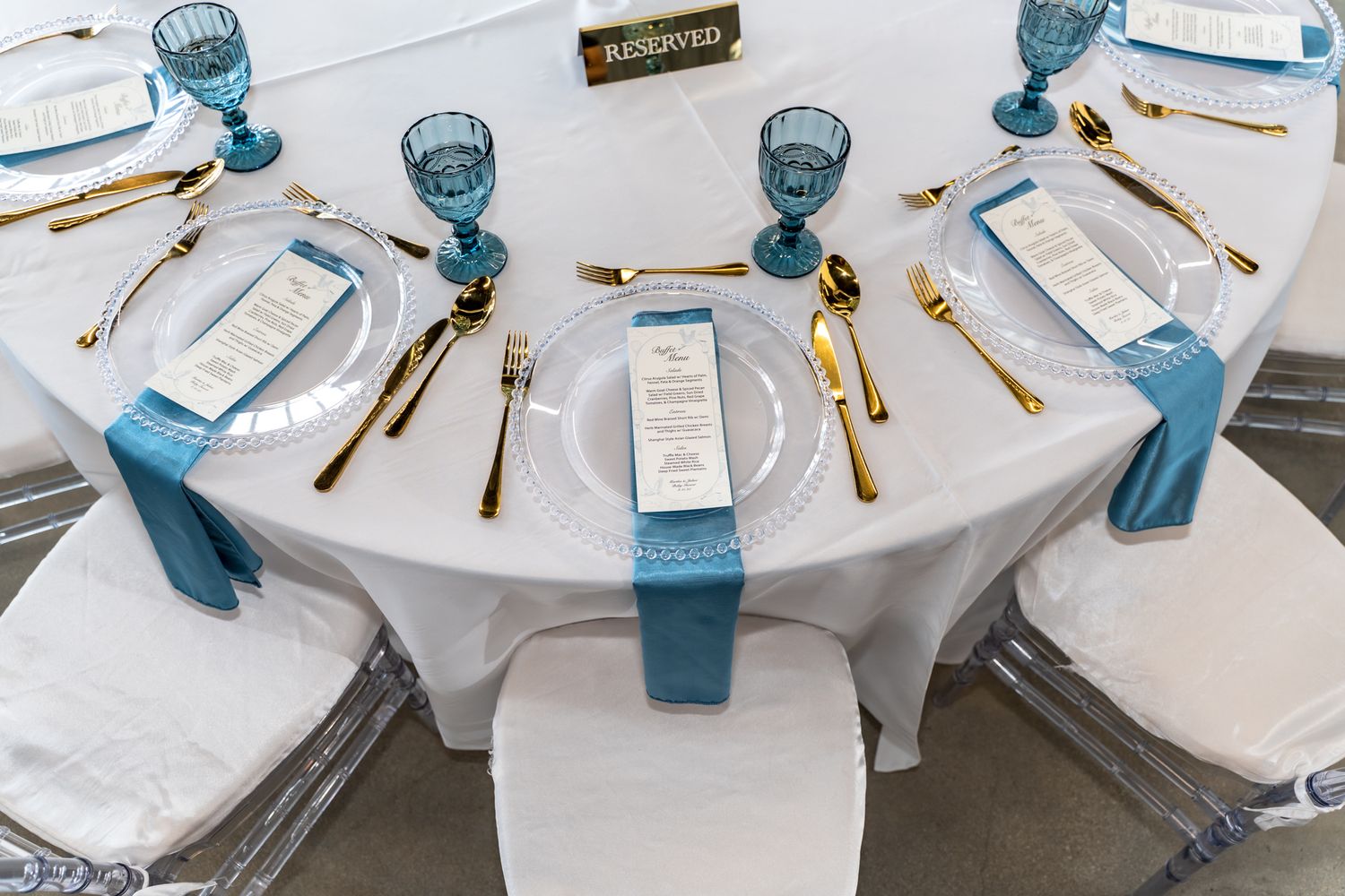 Elegant table setting with blue glassware and gold cutlery on white tablecloth for formal dining event.