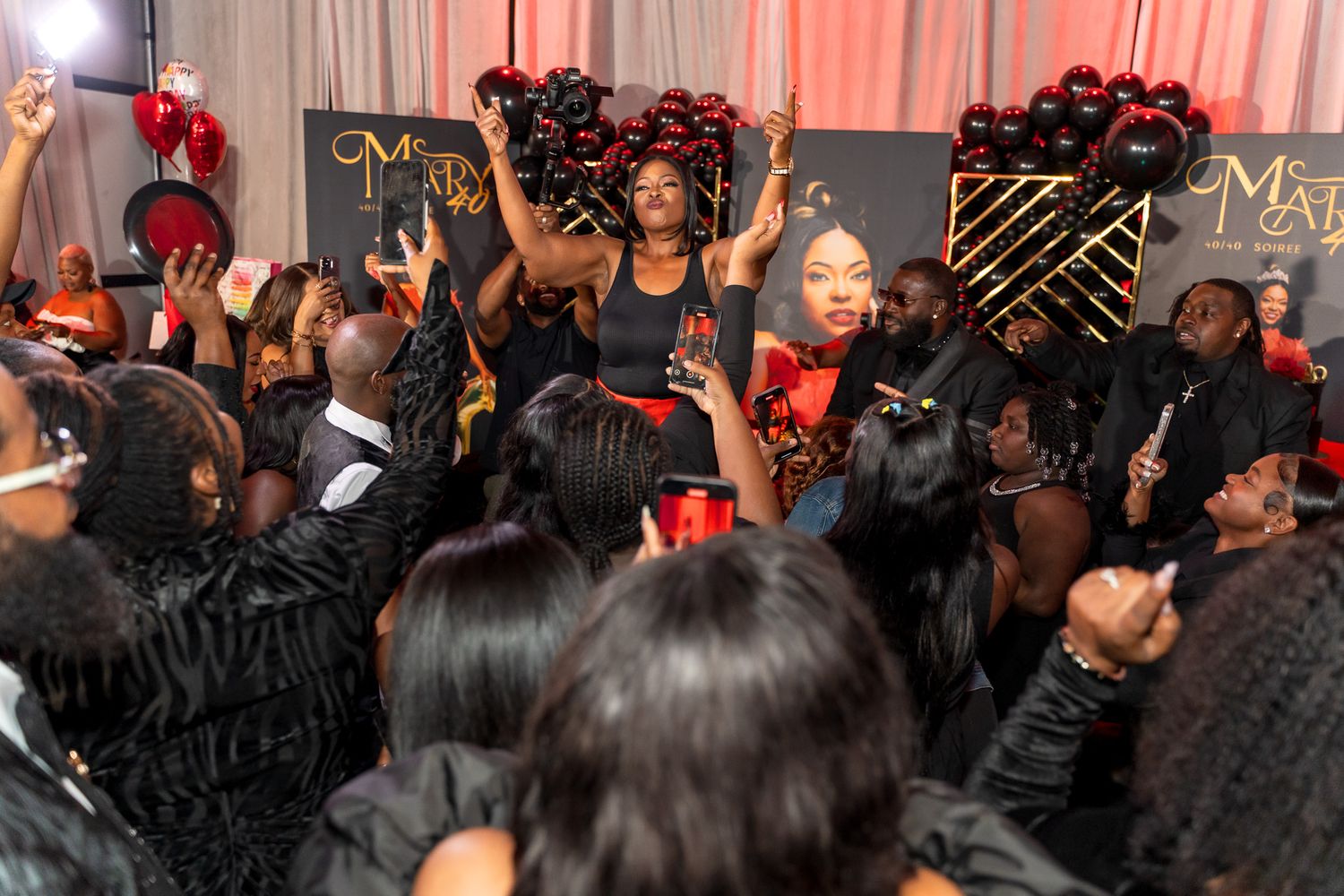 Large group of party guests dancing and celebrating with raised arms in front of decorative black and gold backdrop.