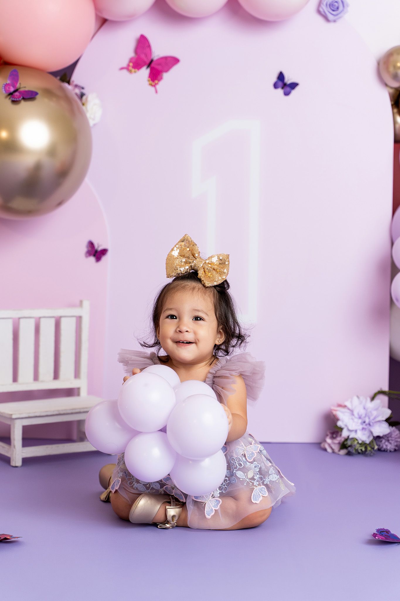 First birthday party setup with pink and purple balloons, butterflies, gold accents and a white decorative chair.
