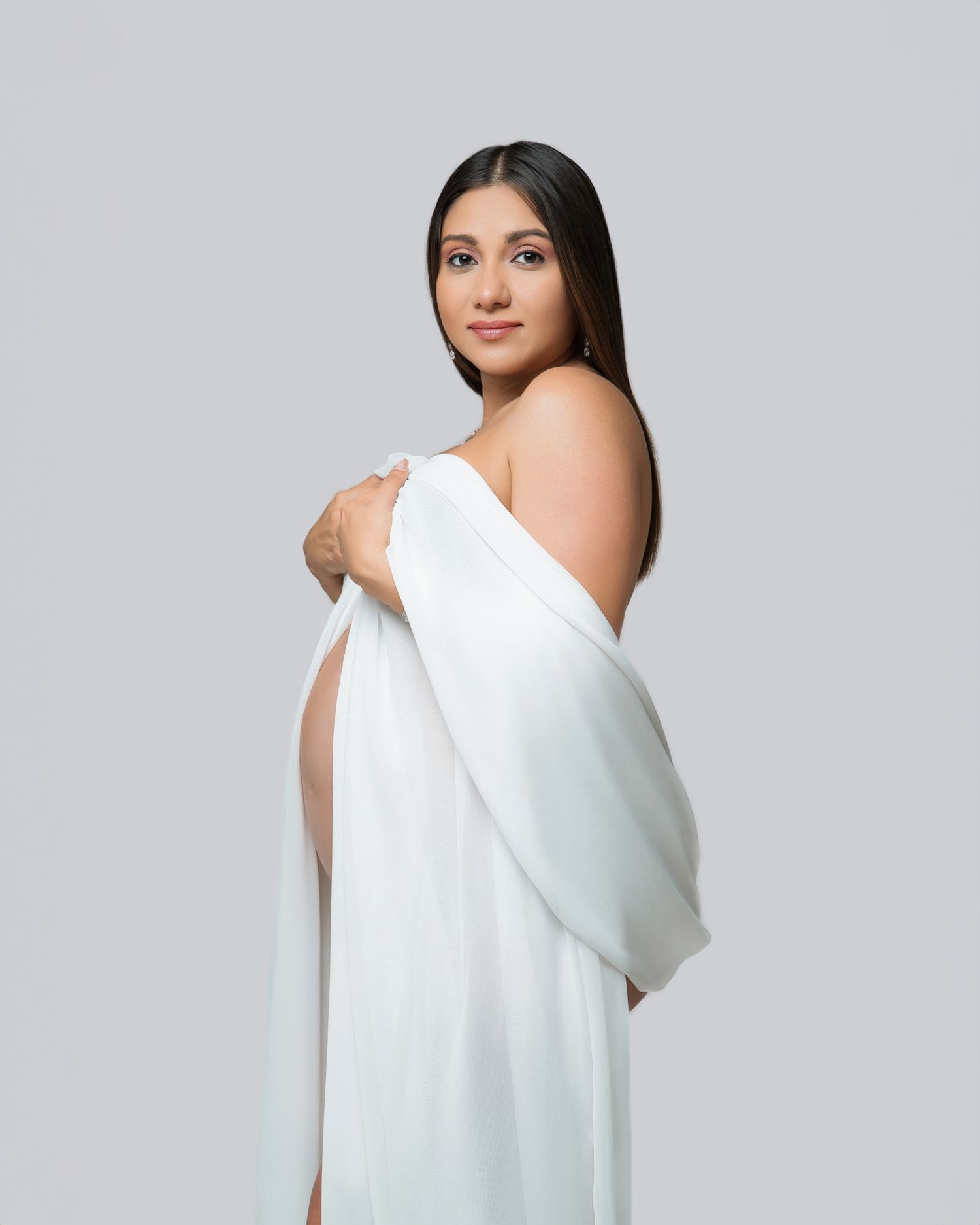 Elegant maternity portrait with figure wrapped in flowing white fabric against grey backdrop.