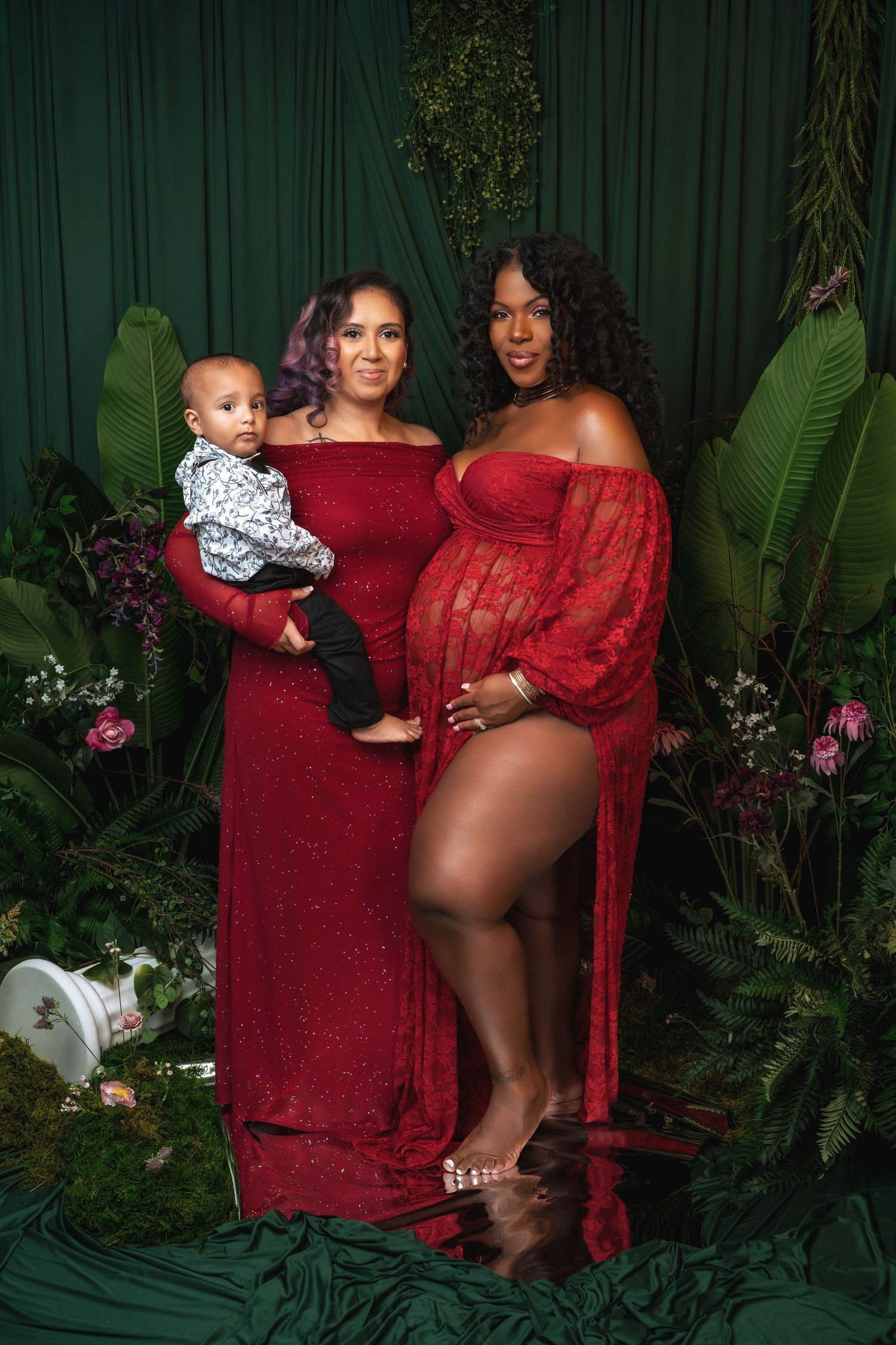 Maternity photoshoot in a tropical garden setting with deep burgundy and red outfits against lush green foliage.