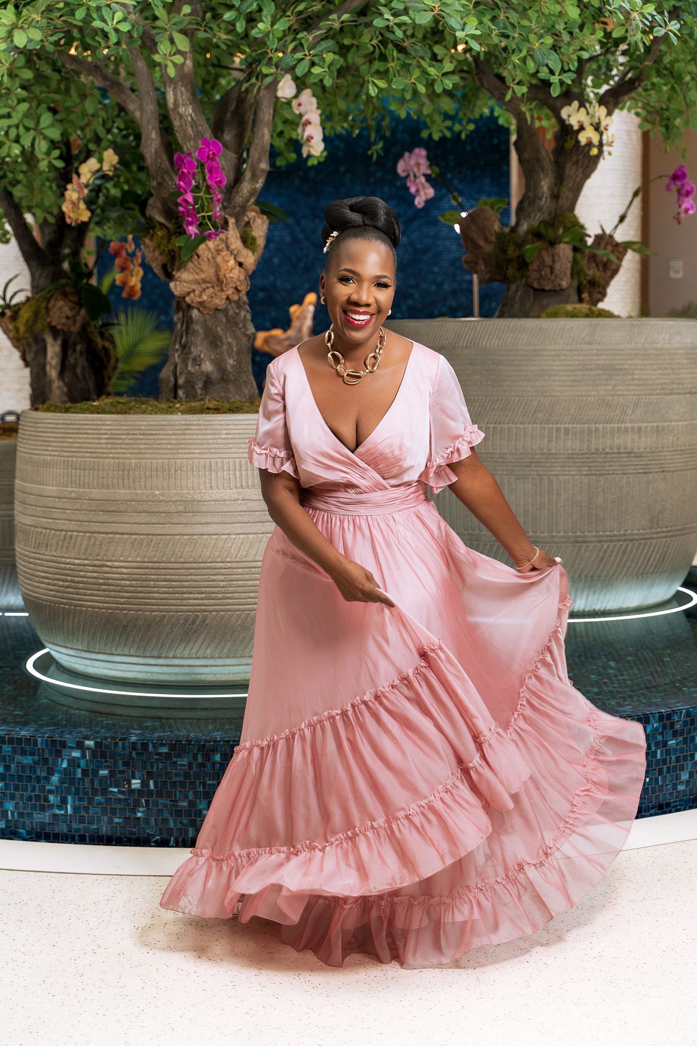 Woman in flowing pink dress twirls near poolside with flowering trees in background.