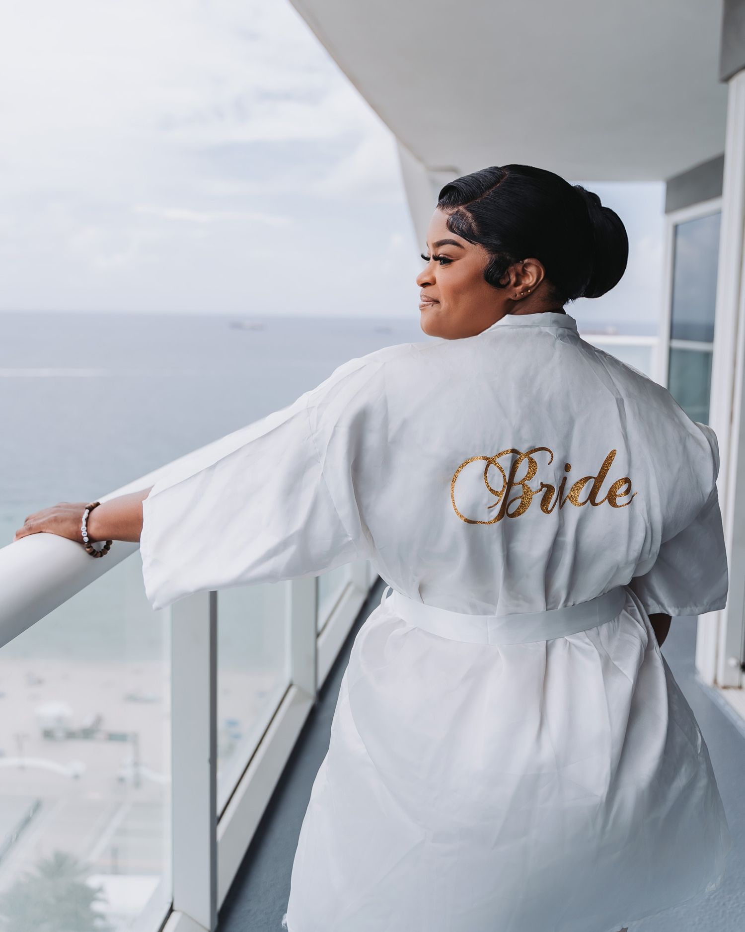 Bride wearing white robe with gold embroidery looking out over balcony view.