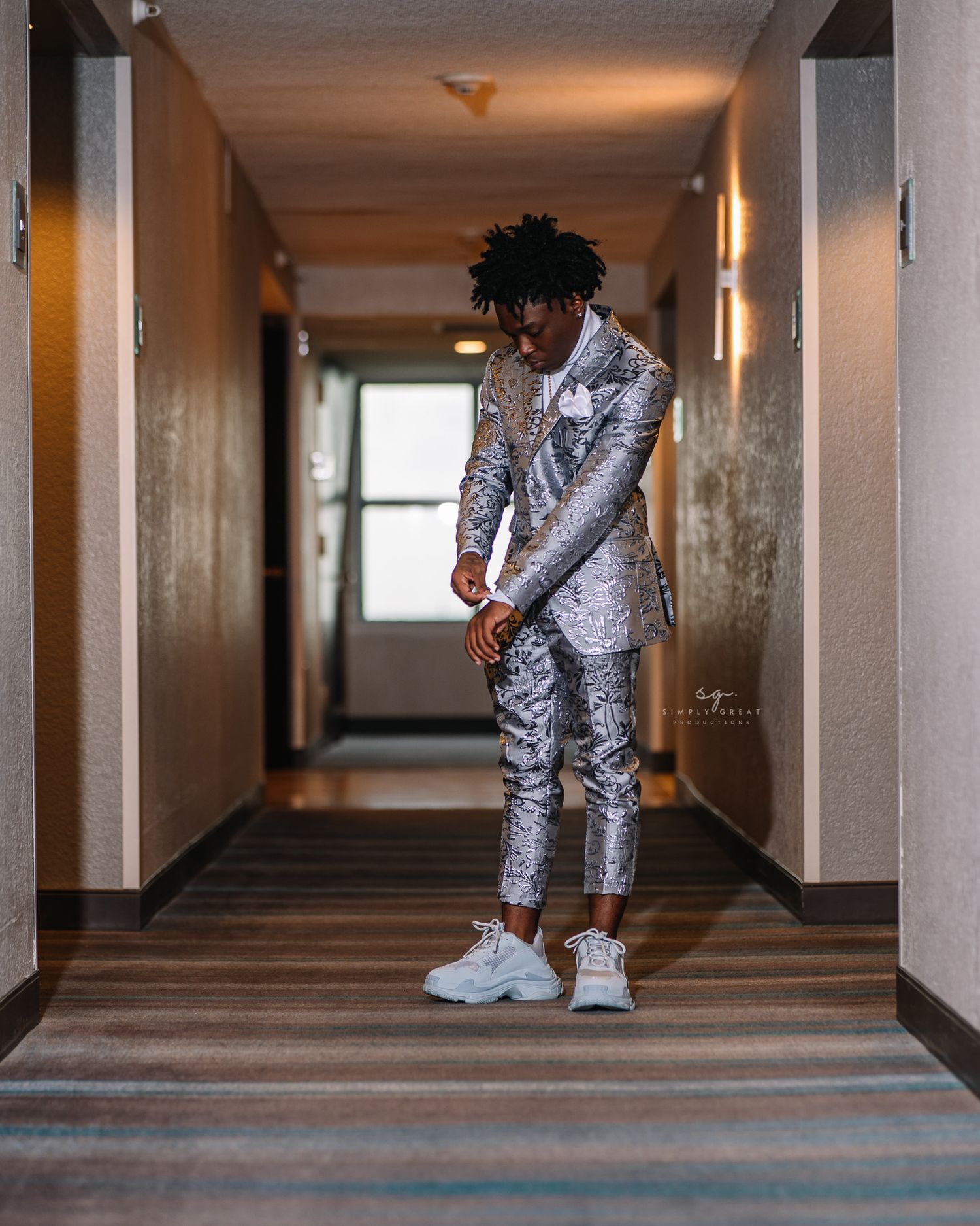 A person wearing a silver patterned suit and white sneakers stands in a hallway.