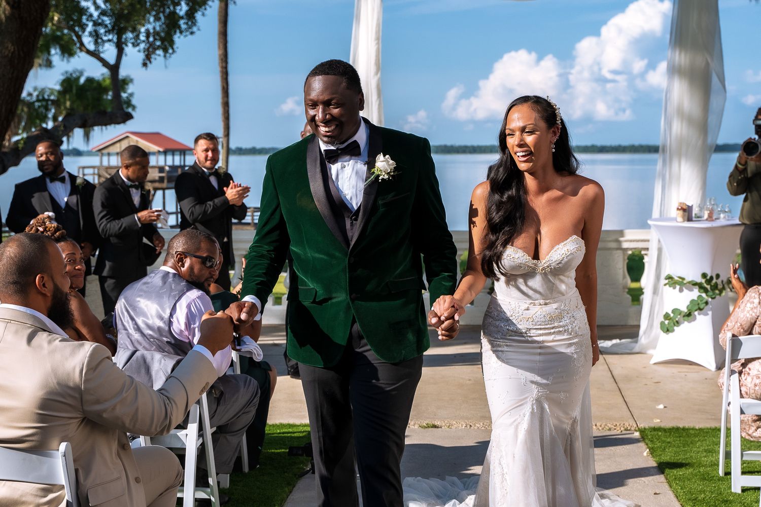 A wedding couple walks down the aisle at an outdoor waterfront ceremony as guests capture the moment.