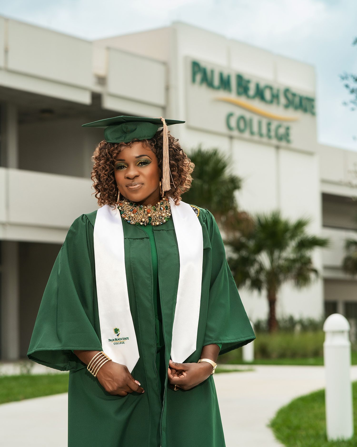 Graduate in traditional green regalia stands outside Palm Beach State College entrance.