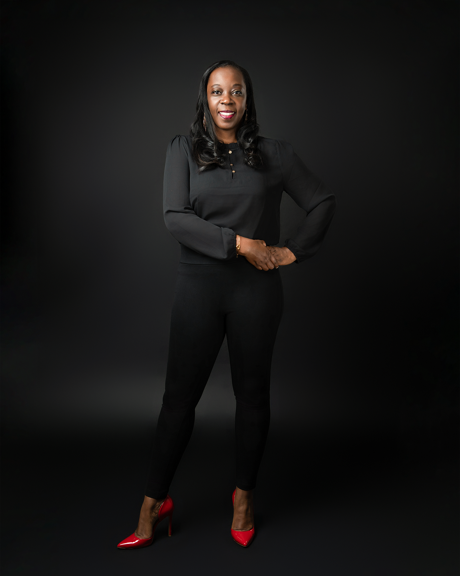 Professional portrait of a person in all black attire with red heels against a dark background.