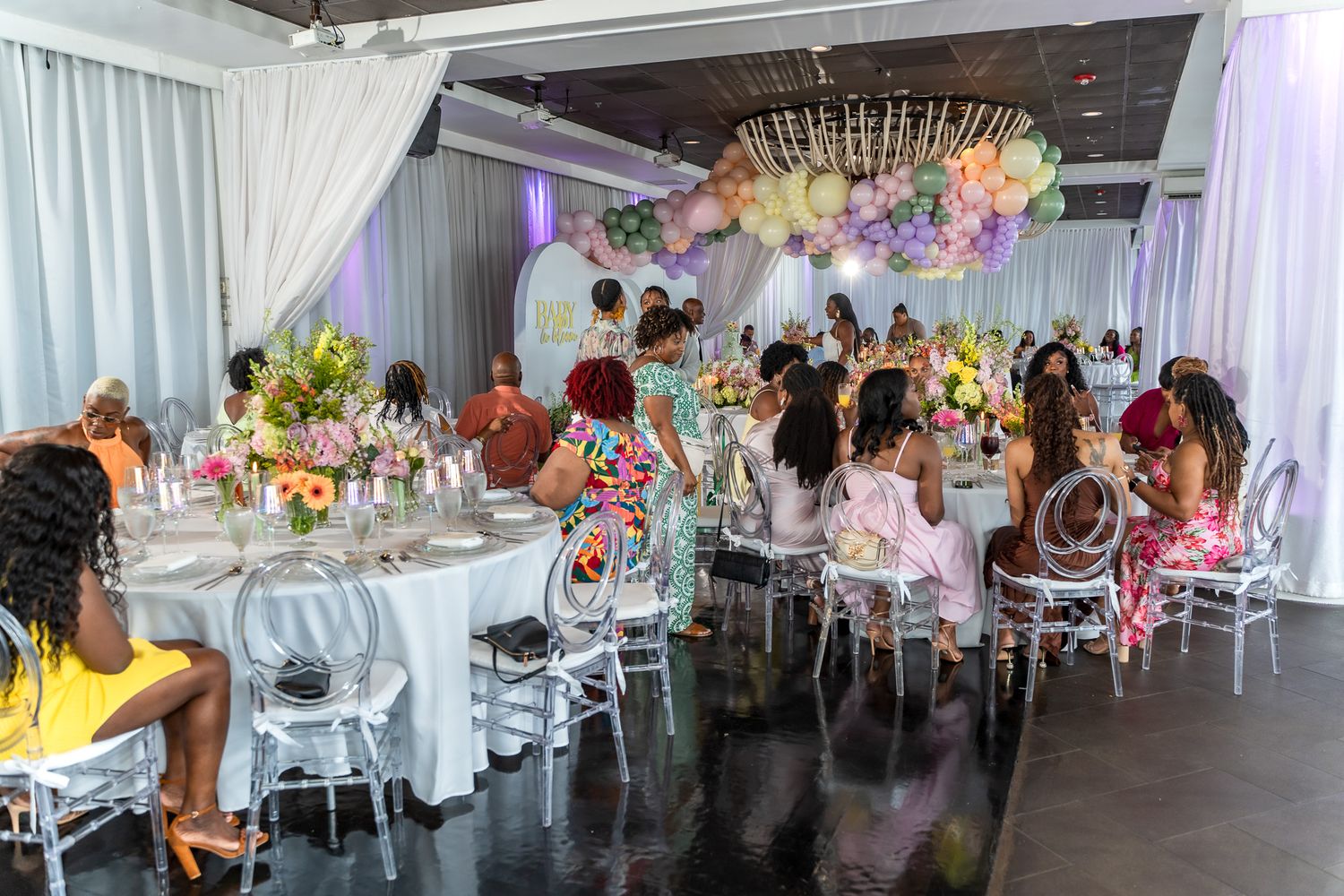 Guests seated at round tables with clear chairs in an elegant event space decorated with balloons and draping.