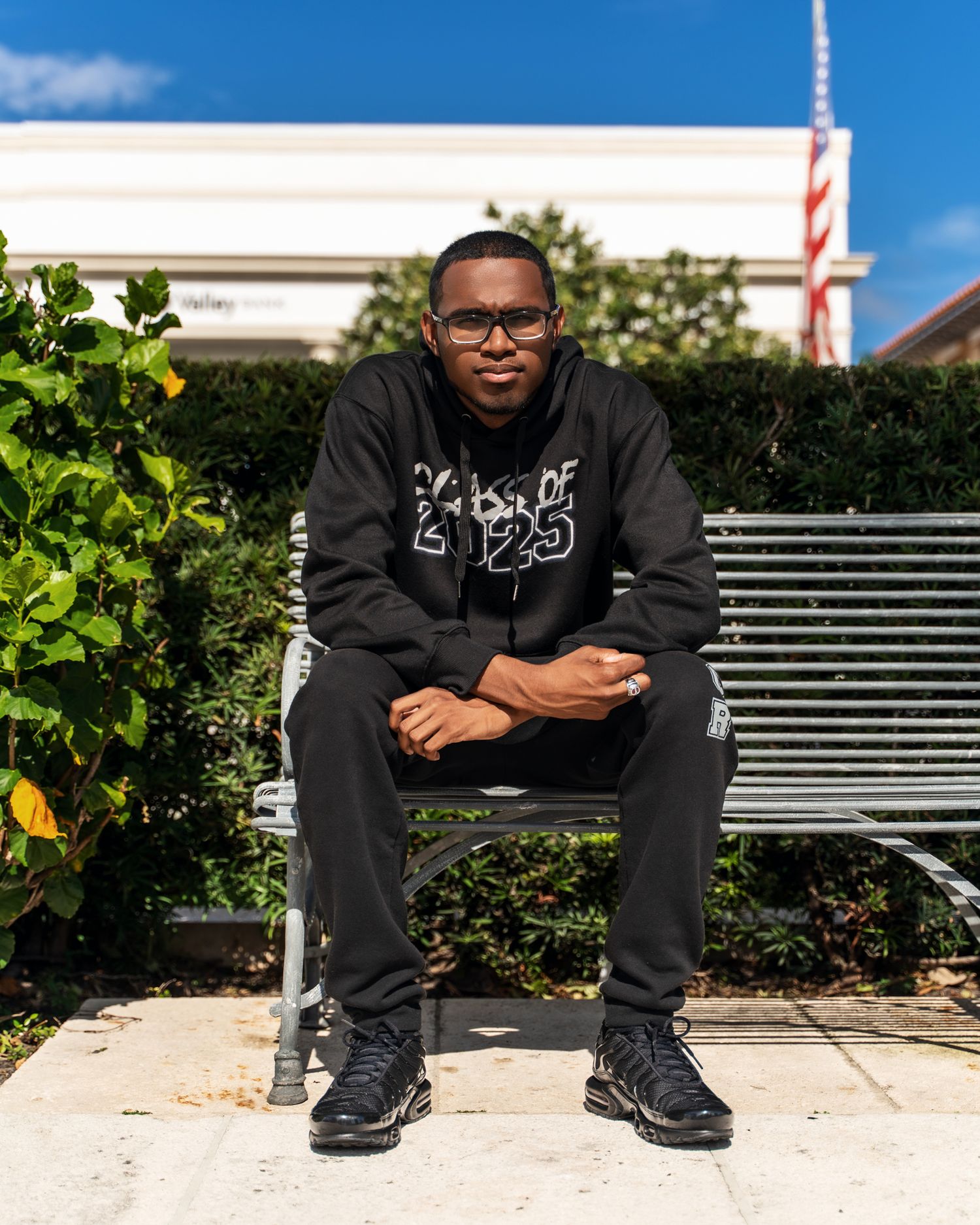 Person in a black sweatshirt and glasses sits casually on a metal bench outdoors on a sunny day.
