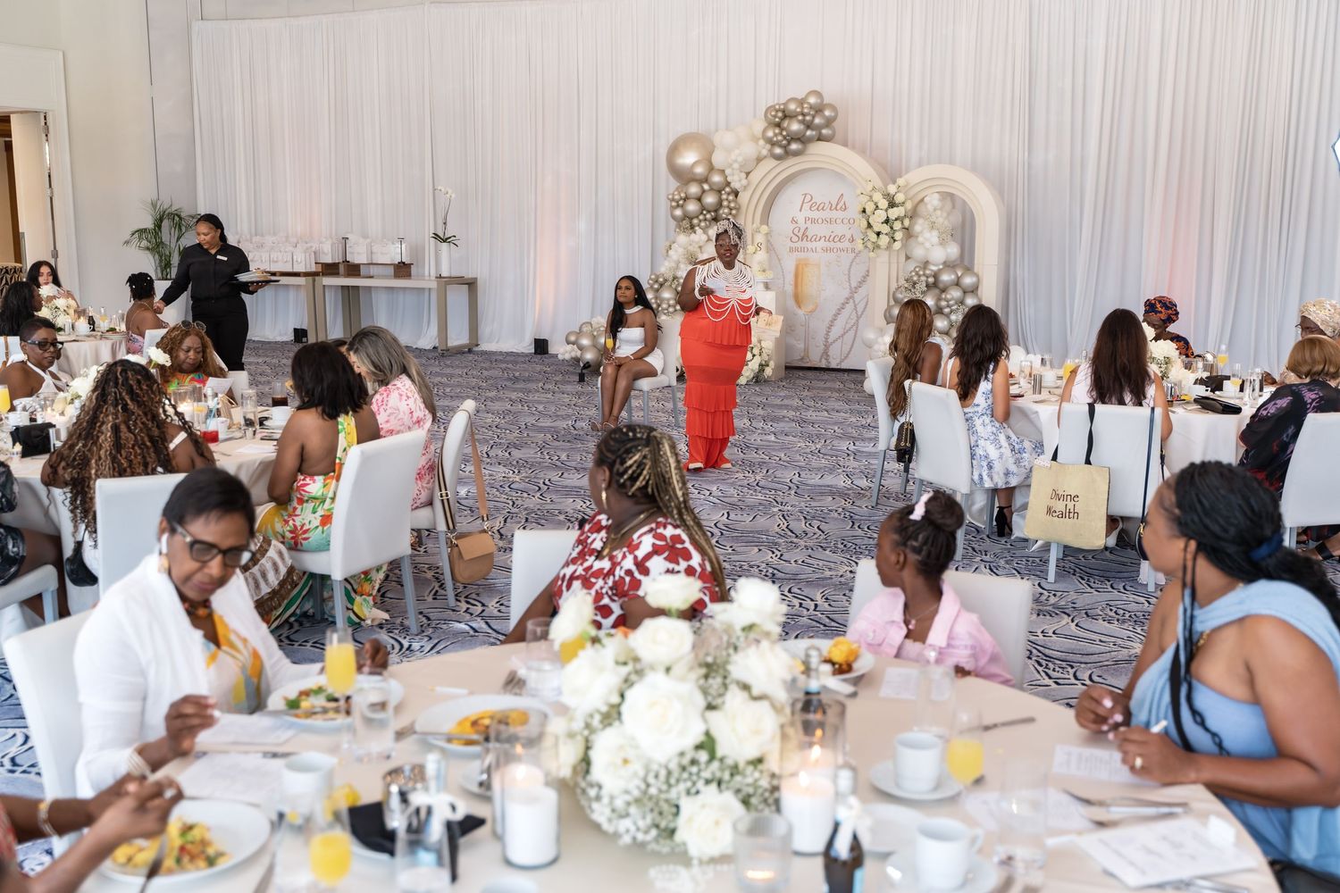 Group of guests enjoying a wedding reception at decorated tables in a light-filled venue with white decor and floral arrangements.