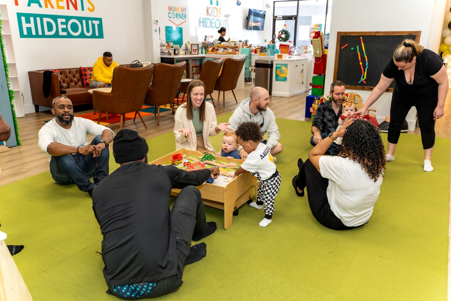Adults and children sitting on green carpet in playroom during social gathering.