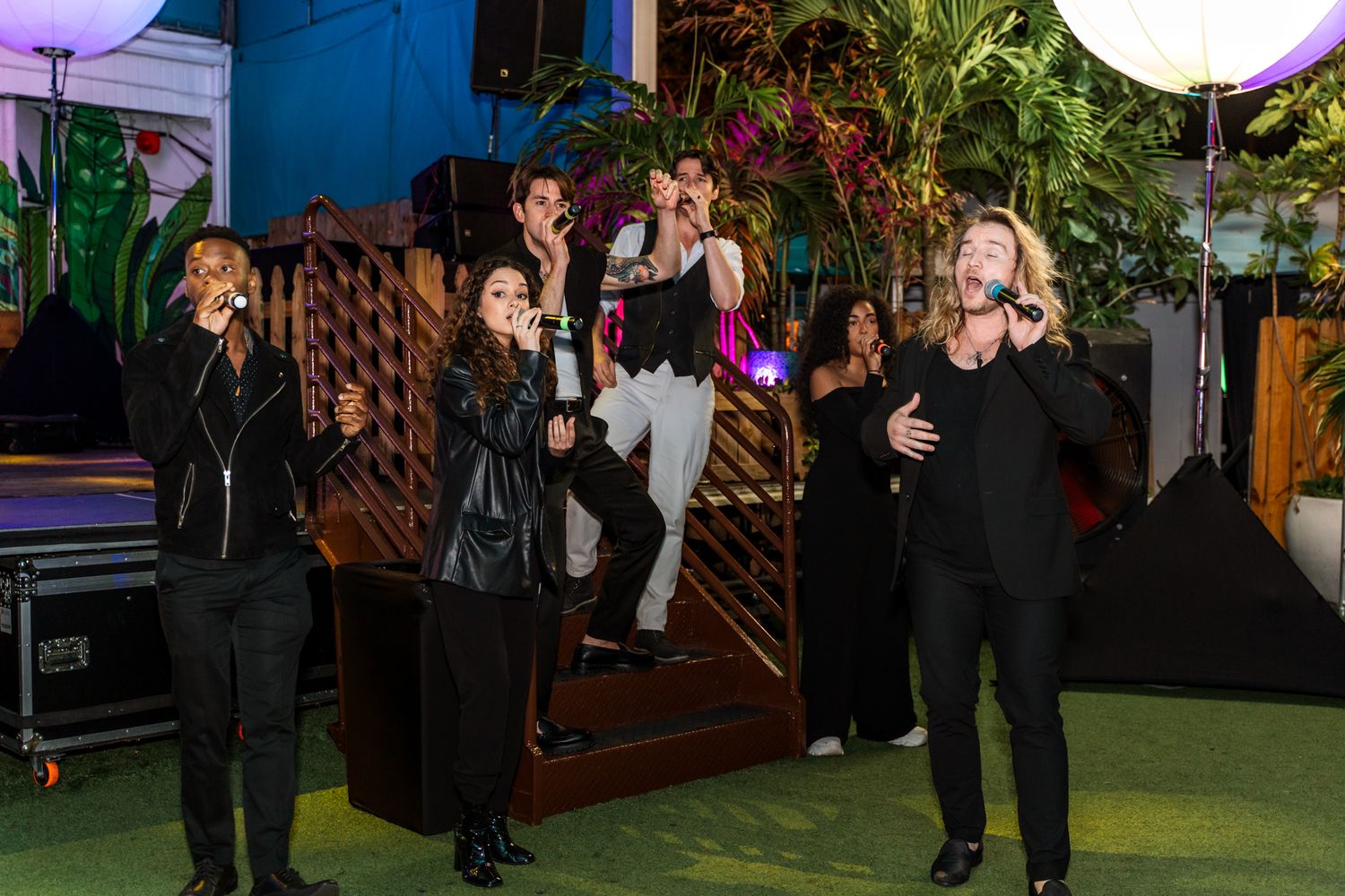 Group performance on outdoor stage with dramatic lighting and wood panel backdrop.