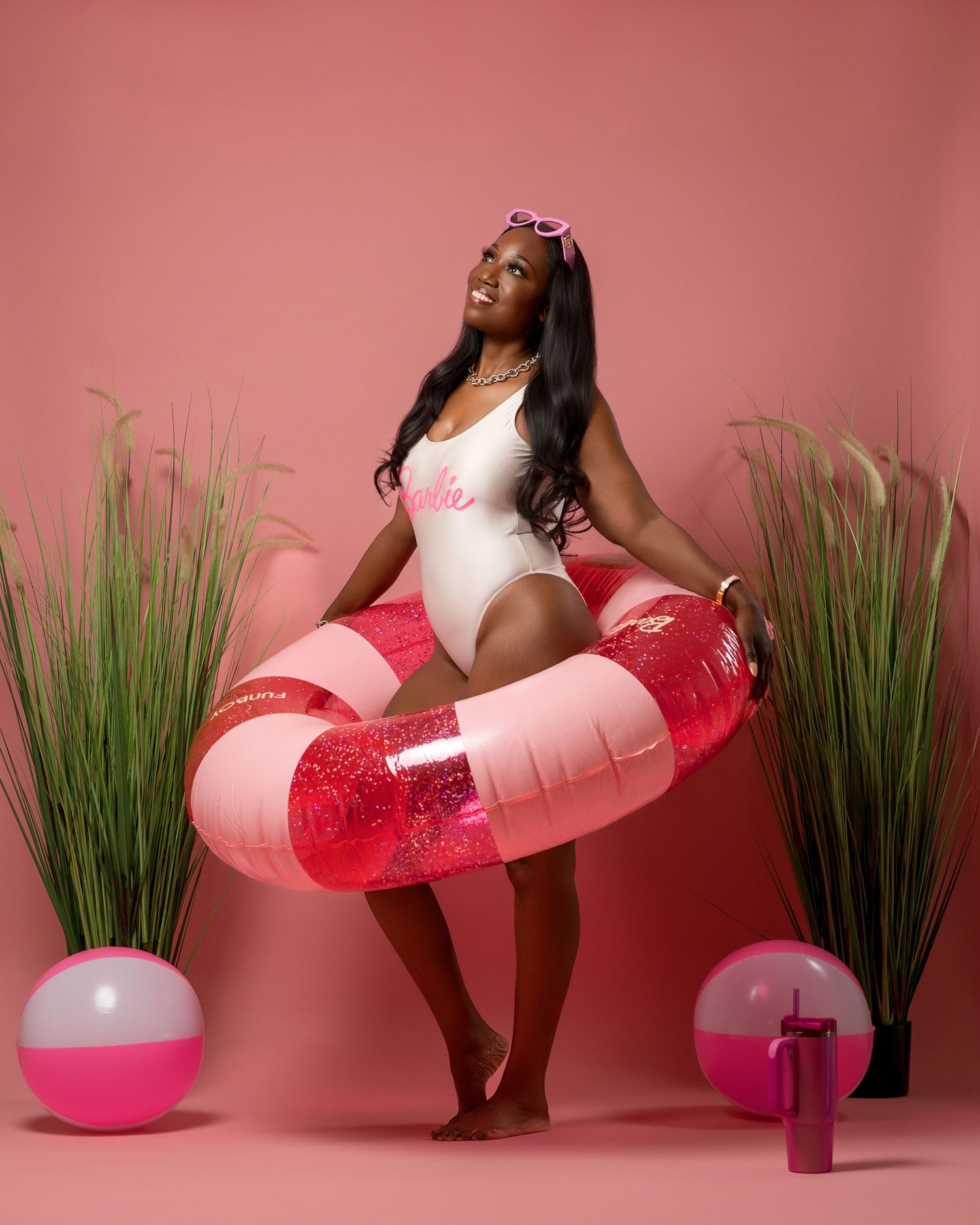 Woman in pink swimsuit poses with glittery pink pool float and beach balls against pink background with decorative grass.