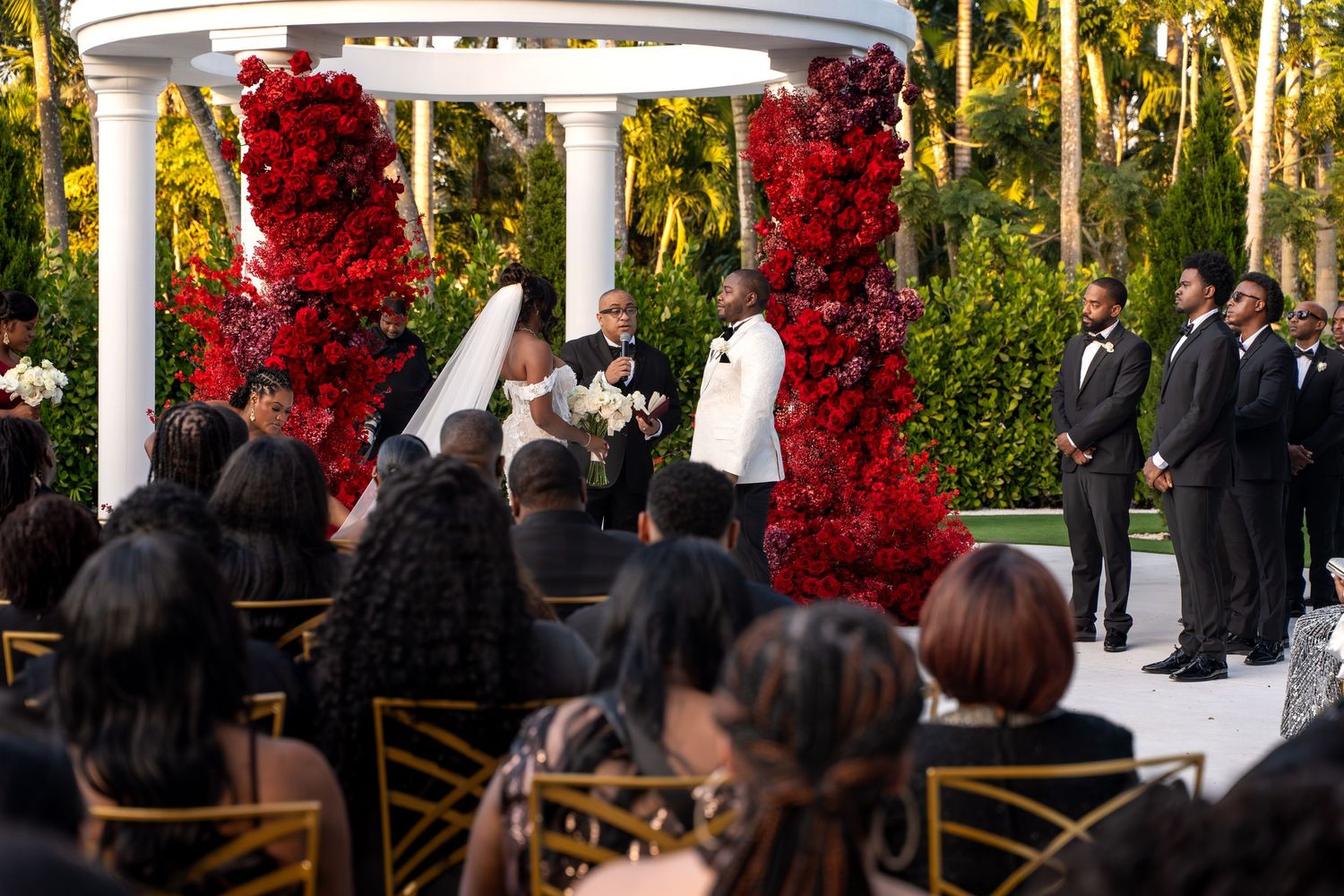 A wedding ceremony setup outdoors with red floral arrangements decorating a white gazebo structure.