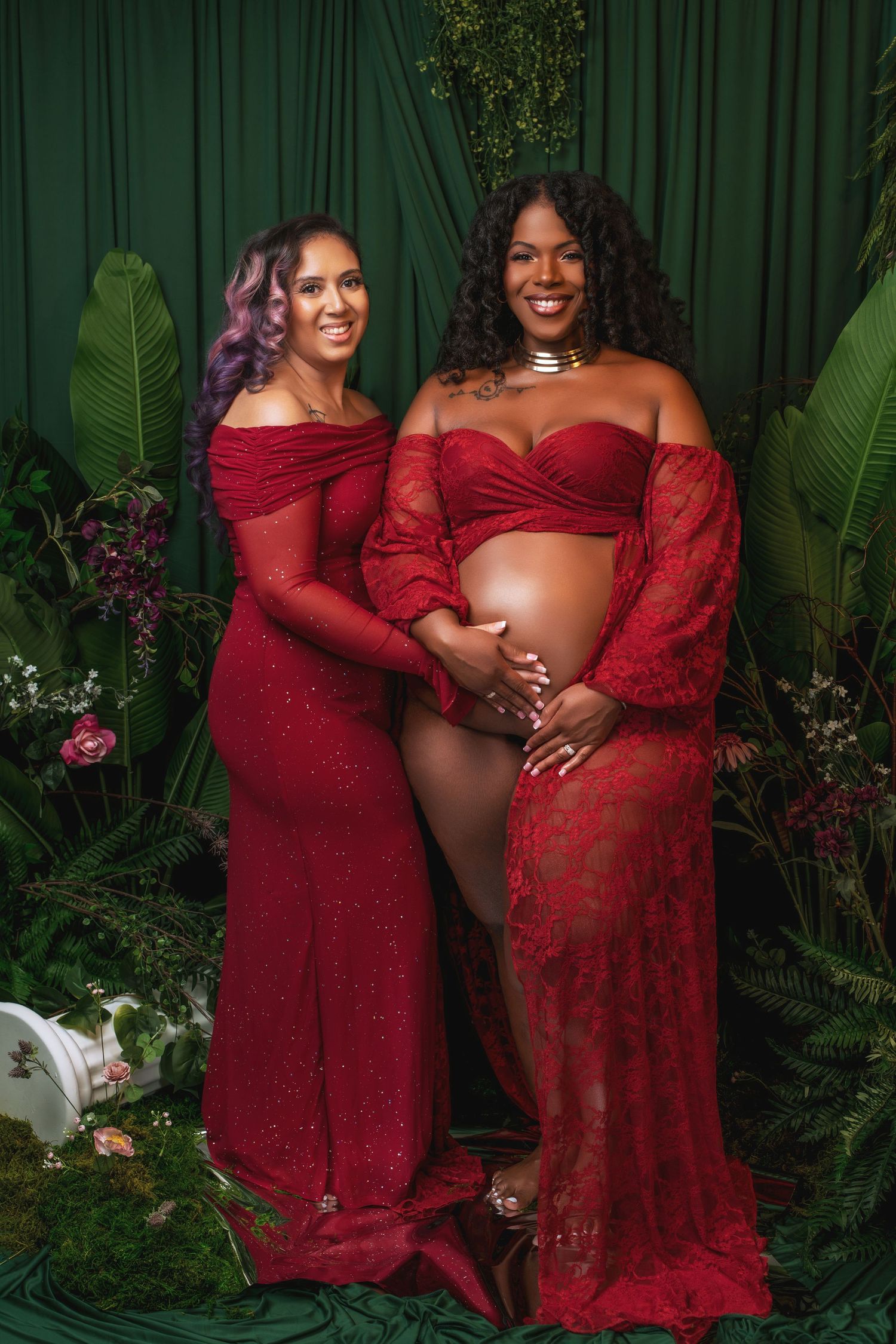 Two people wearing elegant red gowns pose together in a lush garden setting with green foliage and flowers.