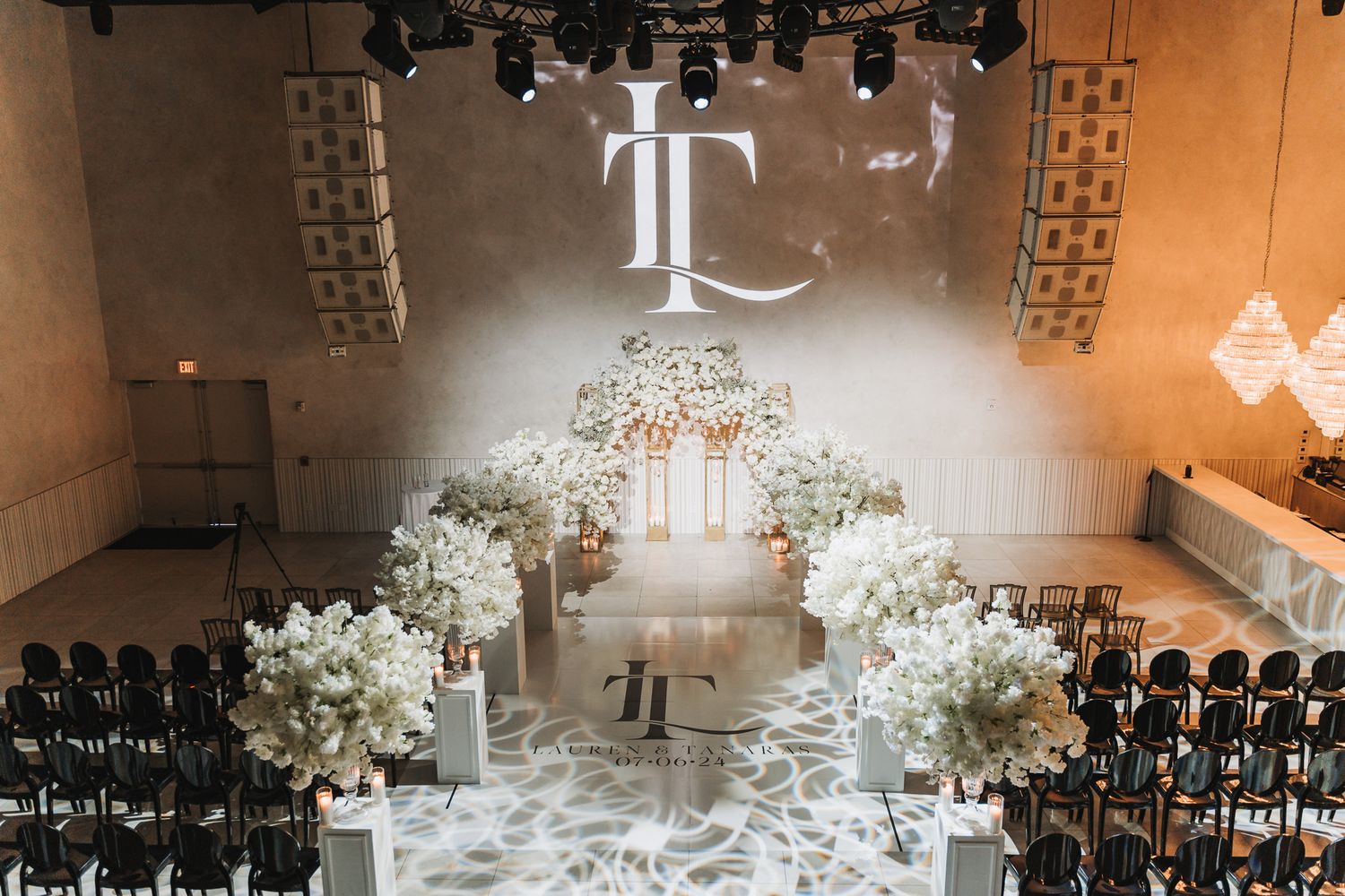 Modern wedding ceremony setup with white floral arrangements and black chairs under a monogram display.