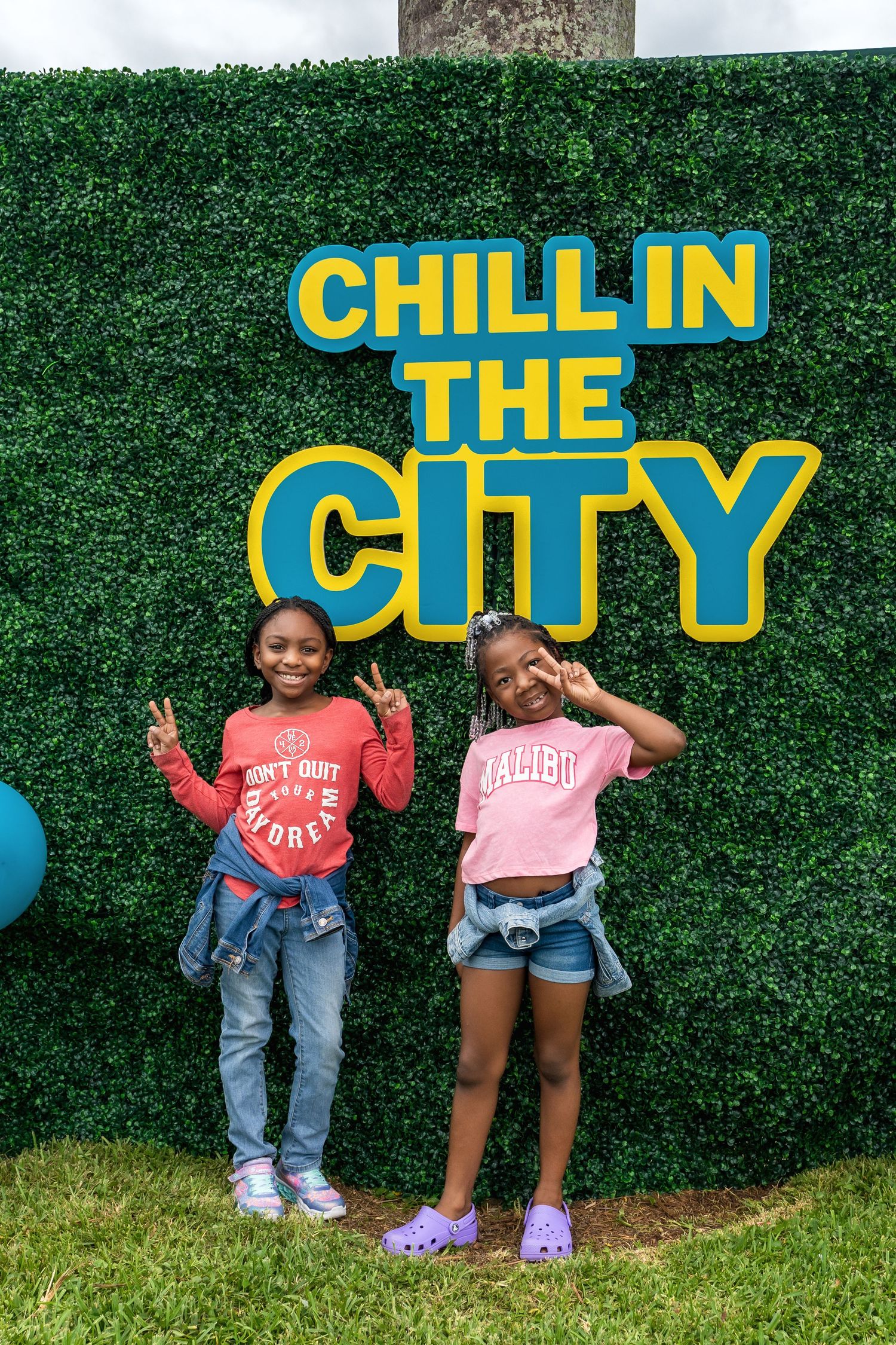Two children posing in front of a green hedge wall with blue and yellow 'Chill in the City' signage.