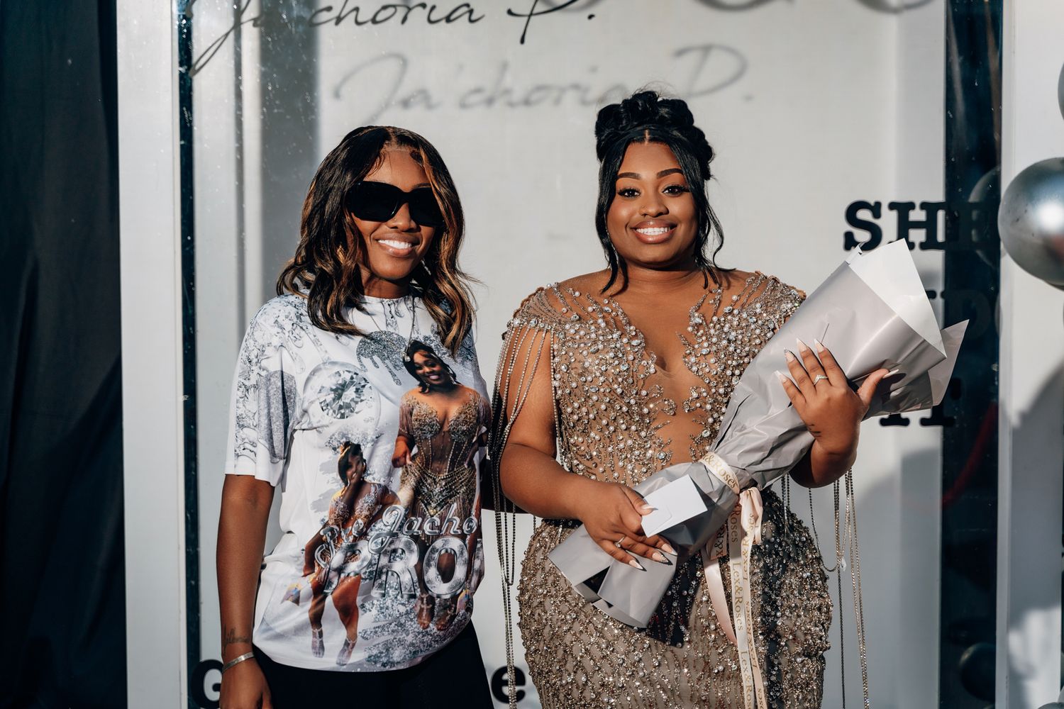 Celebratory photo with graduate in beaded gown and guest in graphic tee holding diplomas.