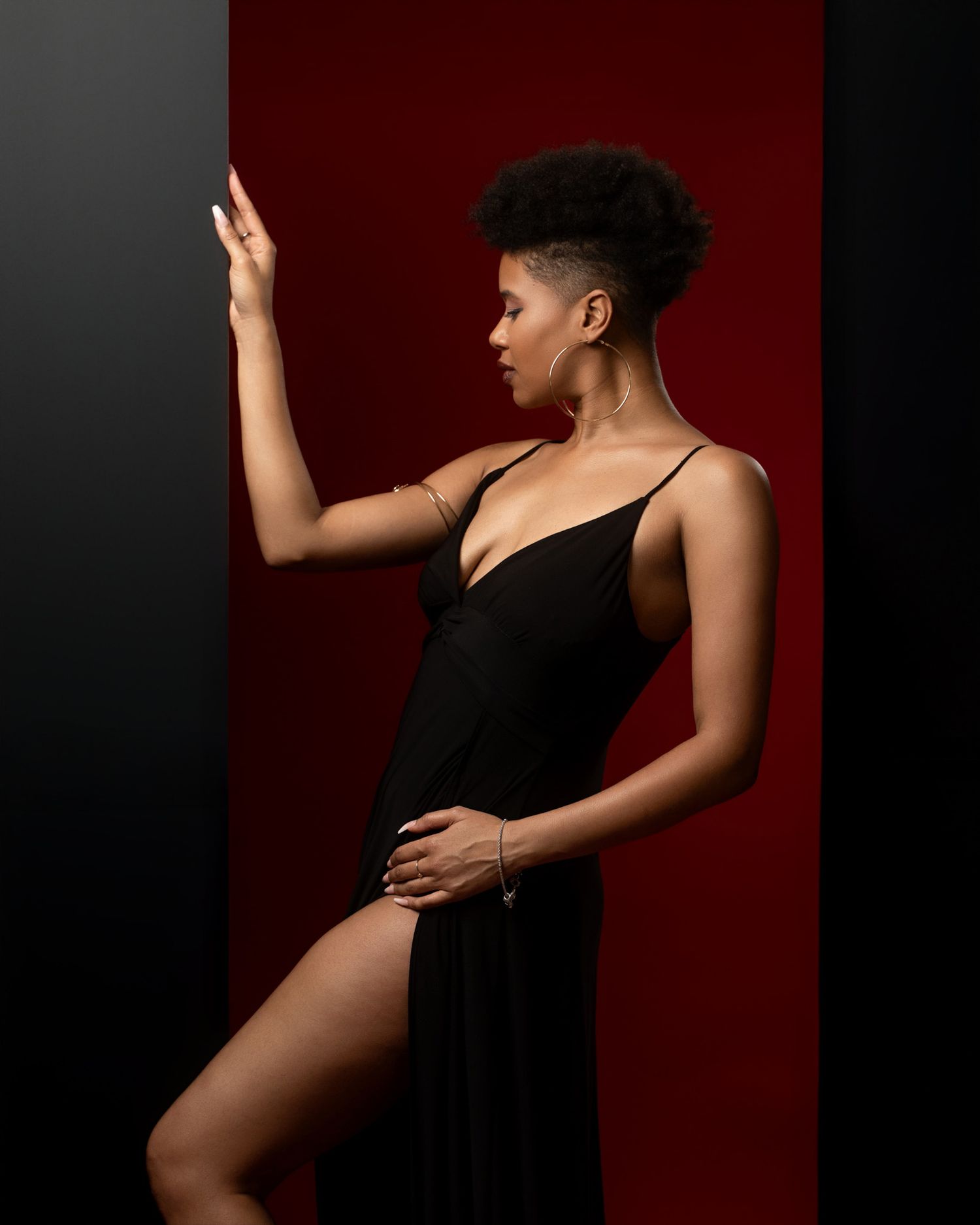 Artistic pose in black evening gown with leg slit against red and black backdrop.