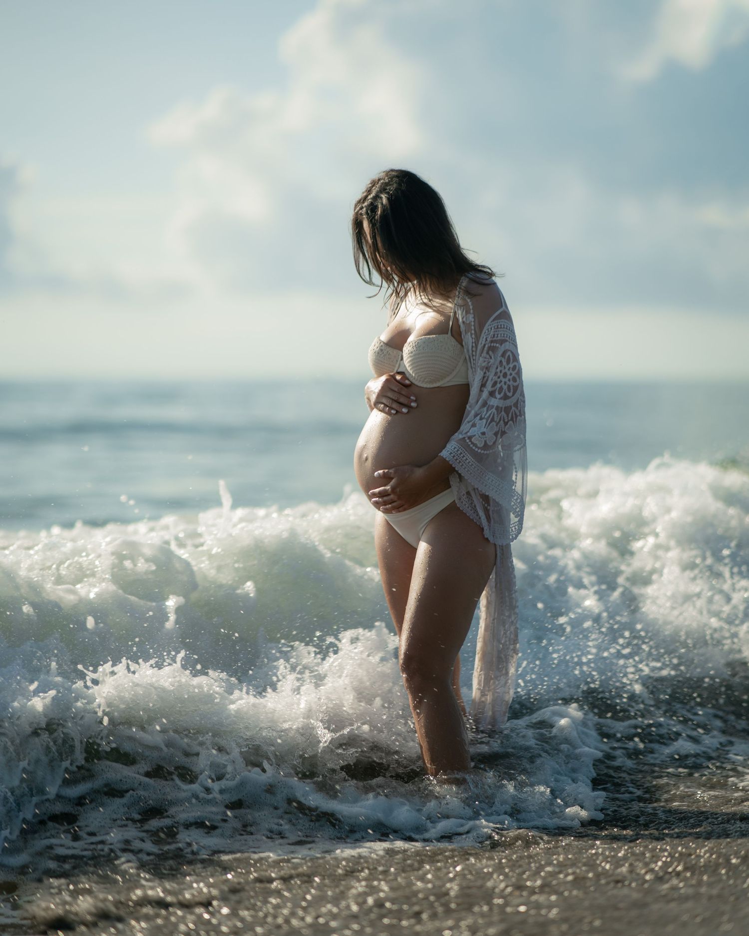 Pregnant woman in white swimwear standing in ocean waves during sunset photoshoot.