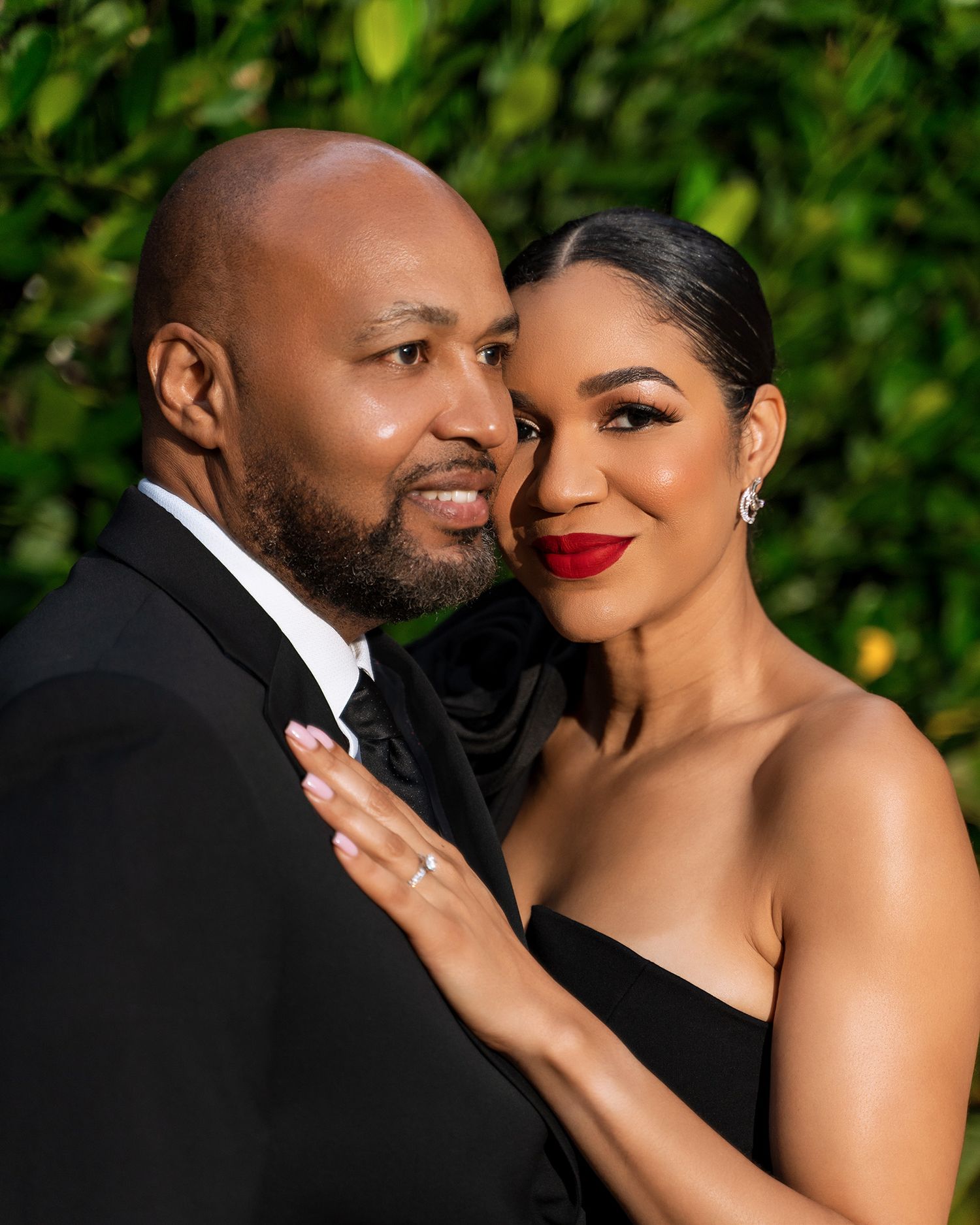 Elegant couple in formal attire pose against green foliage background.