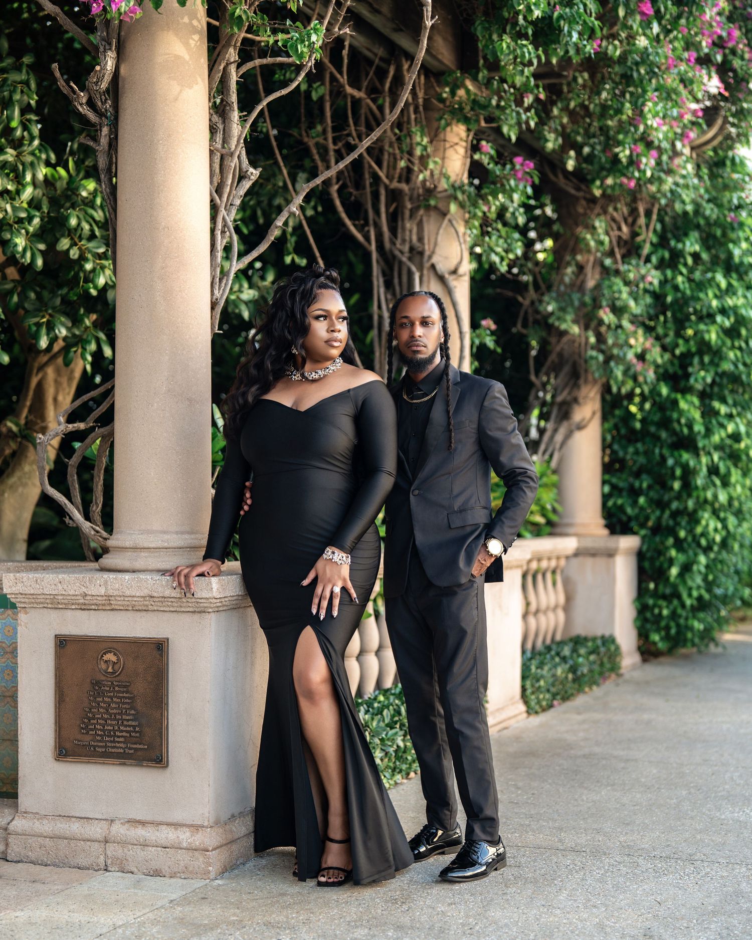 Elegant couple poses together in formal attire by ornate stone columns with flowering vines and greenery in background.
