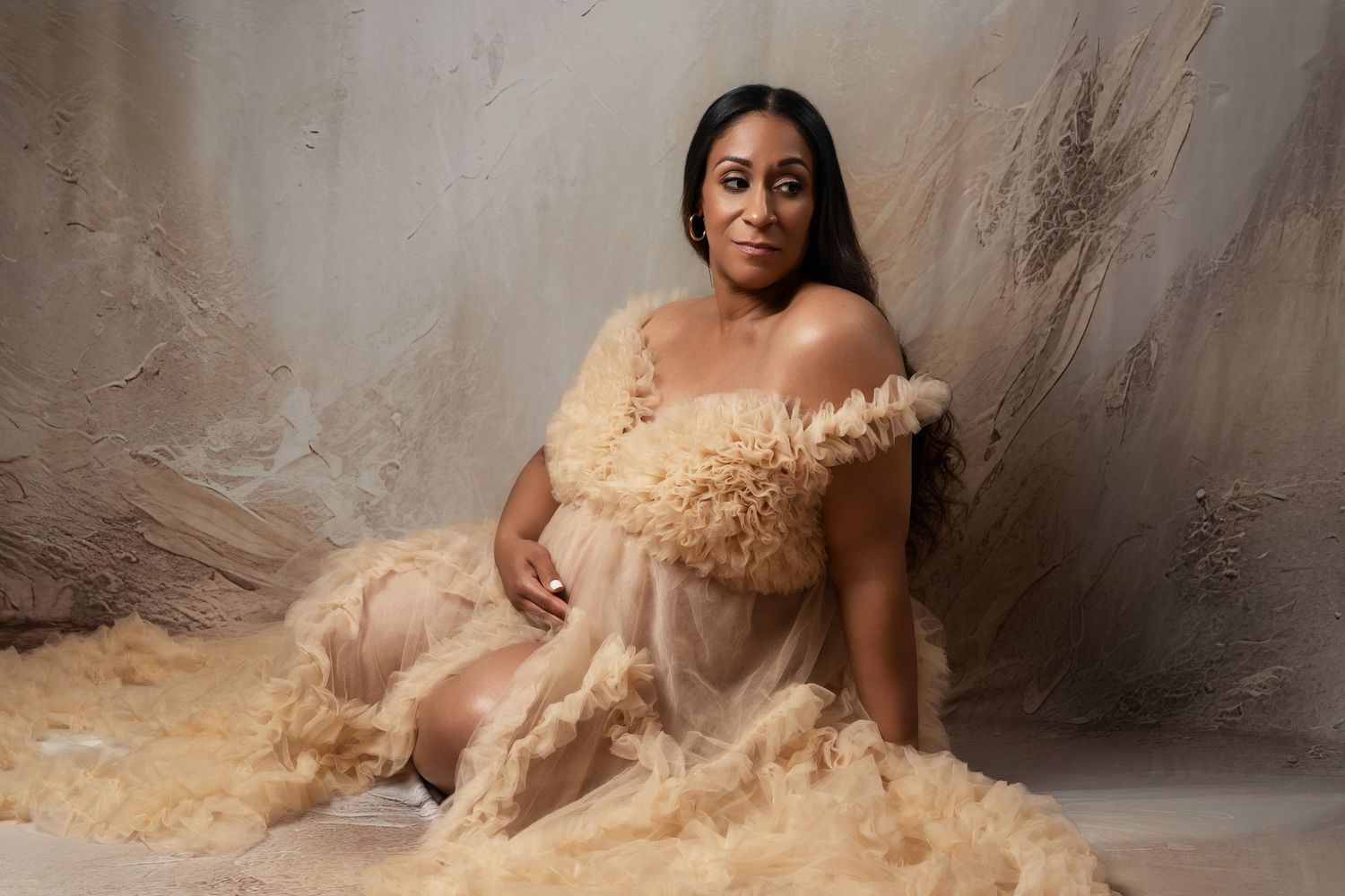 A person wearing a flowing beige tulle gown poses elegantly against a textured gray backdrop.