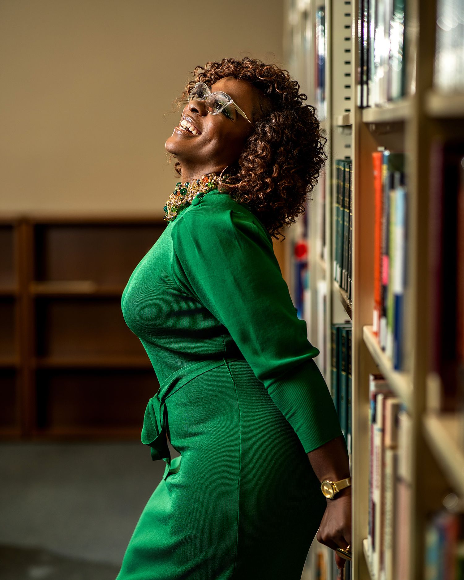 Graduate student in emerald green dress poses confidently near library bookshelves.