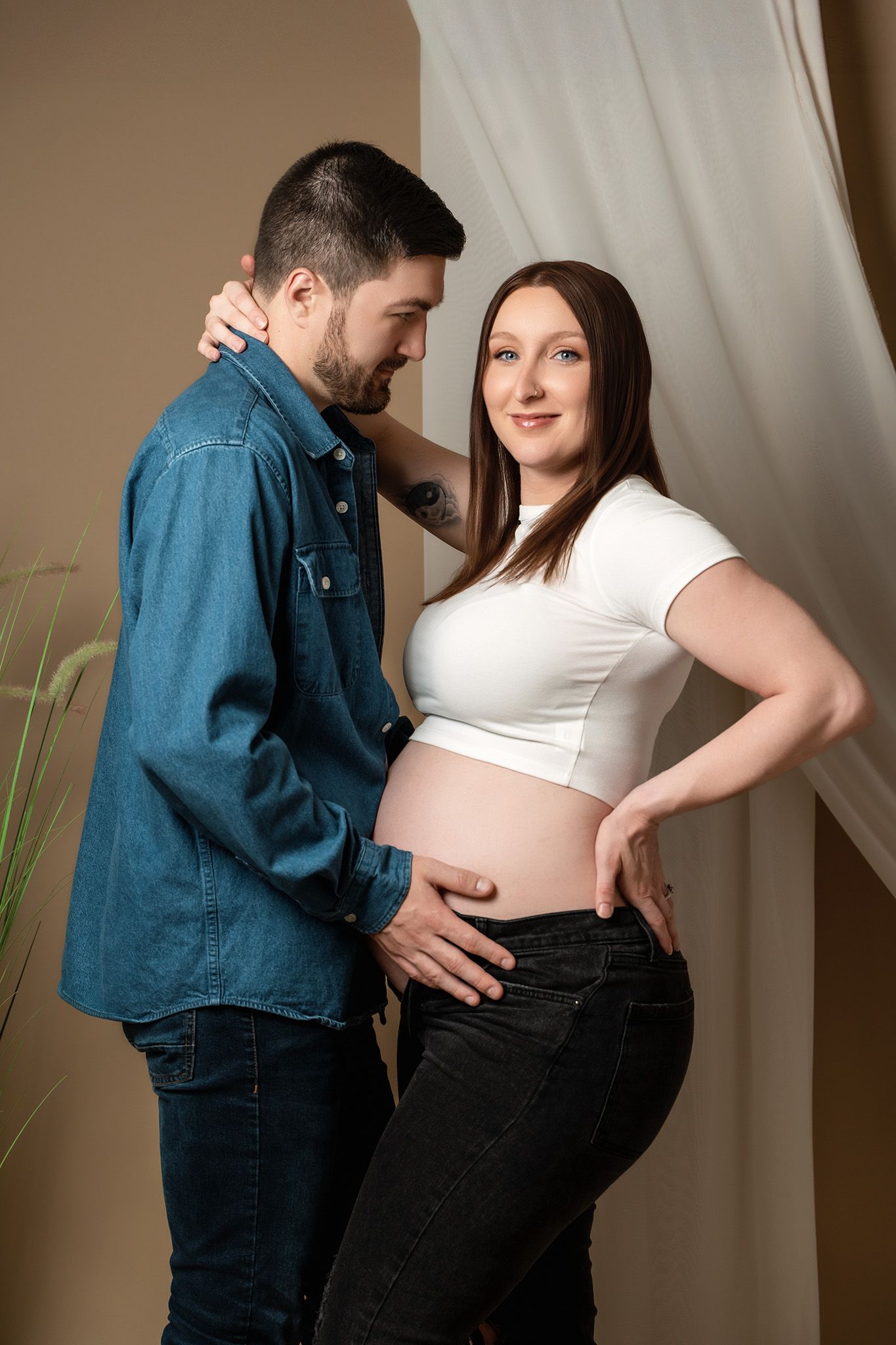 Intimate maternity photo with couple in casual attire against cream curtain and plant decor.