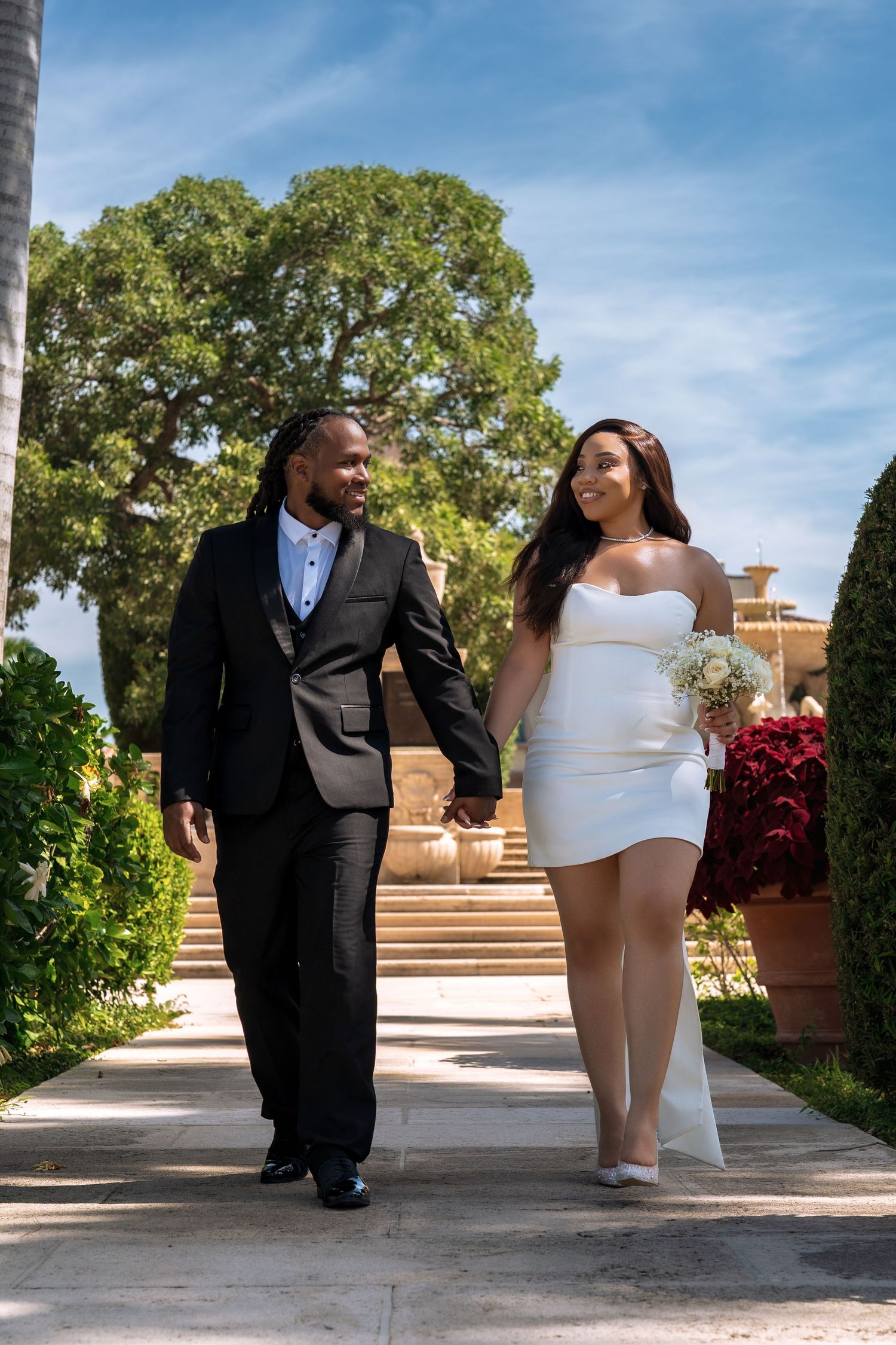 A couple in formal wedding attire walking hand in hand along a sunny garden path.