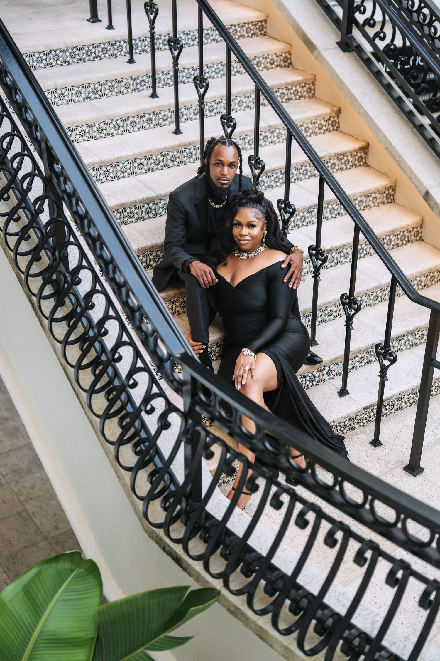 A couple in black formal wear poses for a romantic portrait on an ornate staircase with decorative railings.