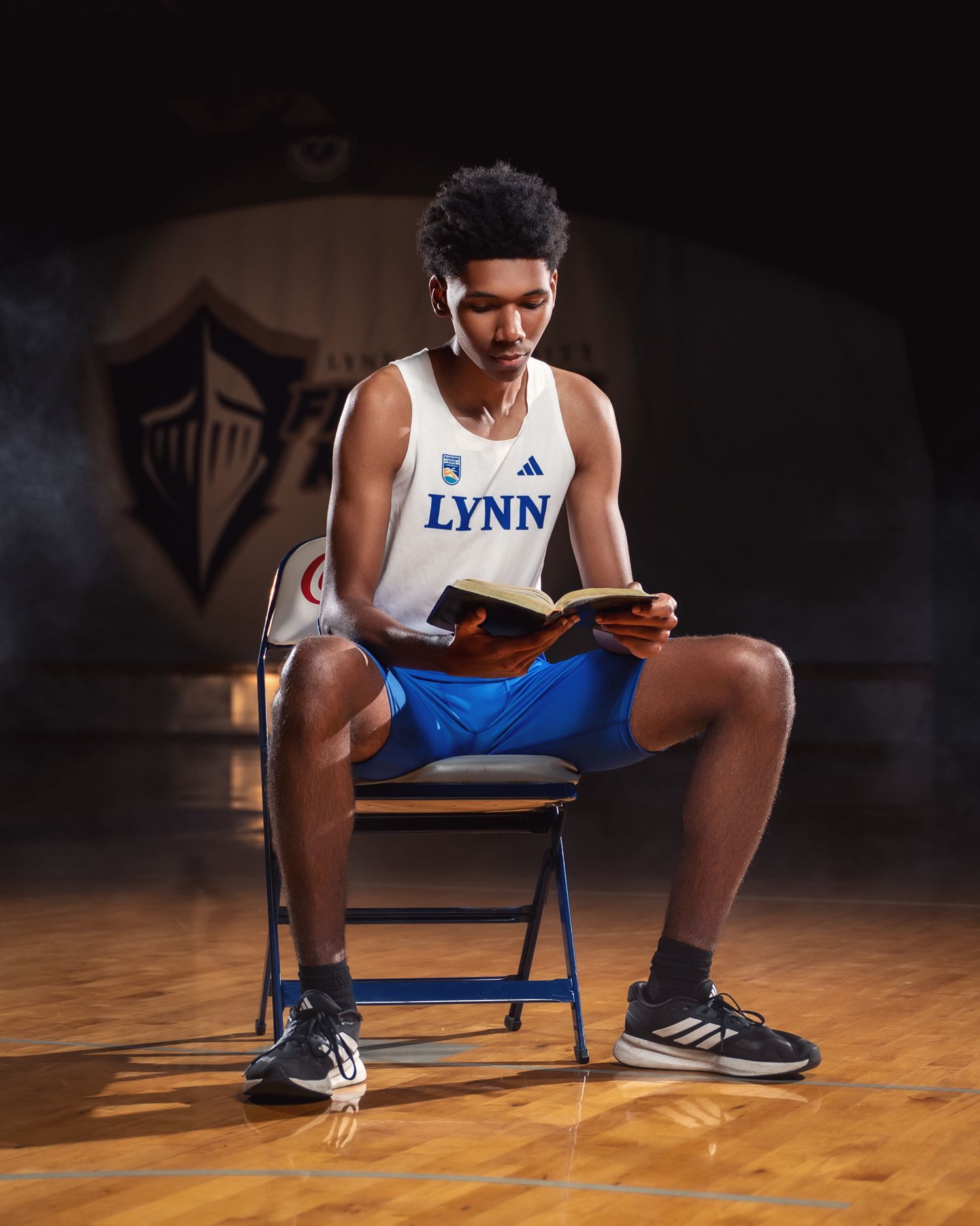 Athletic student in Lynn University uniform sits reading on a blue folding chair in a gymnasium.
