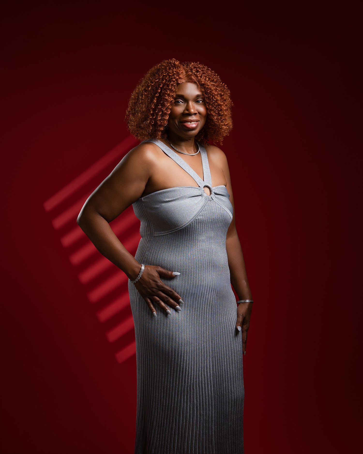Portrait against red backdrop showing grey dress and vibrant curly hair.