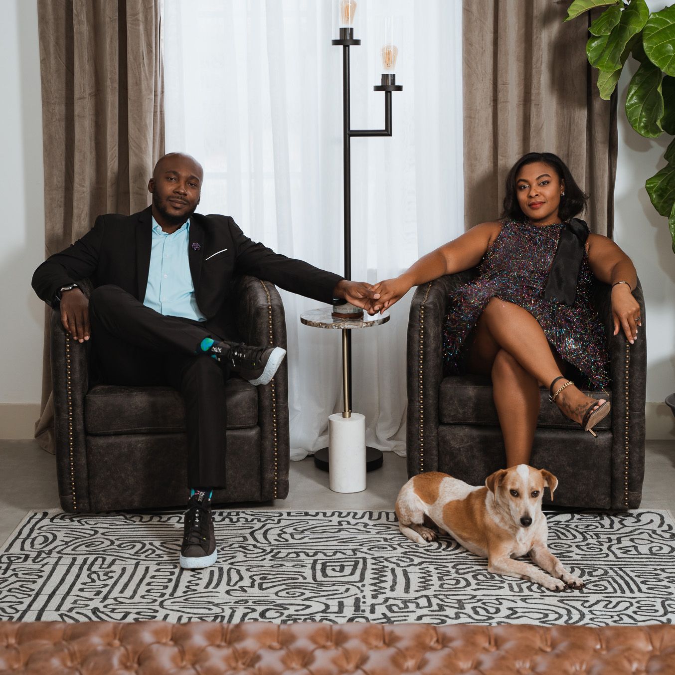 Couple holding hands while seated in armchairs with dog resting on patterned rug.