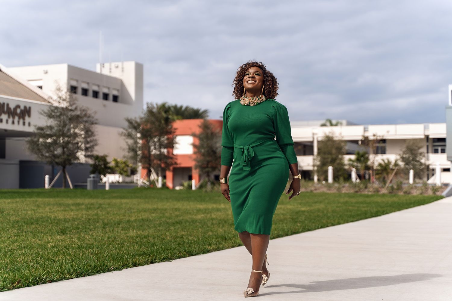 Figure in green dress walks along concrete path with buildings in background.