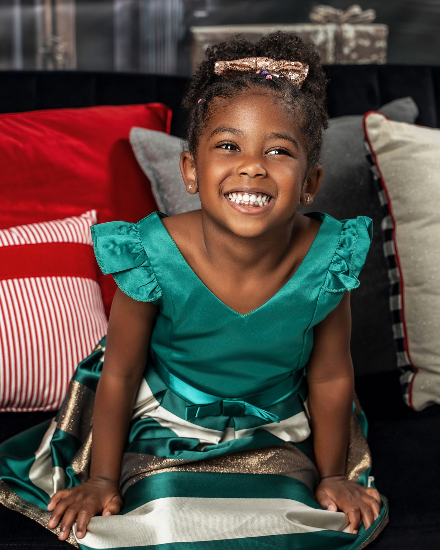 A joyful child wearing a ruffled emerald green holiday dress poses on a couch with red pillows.