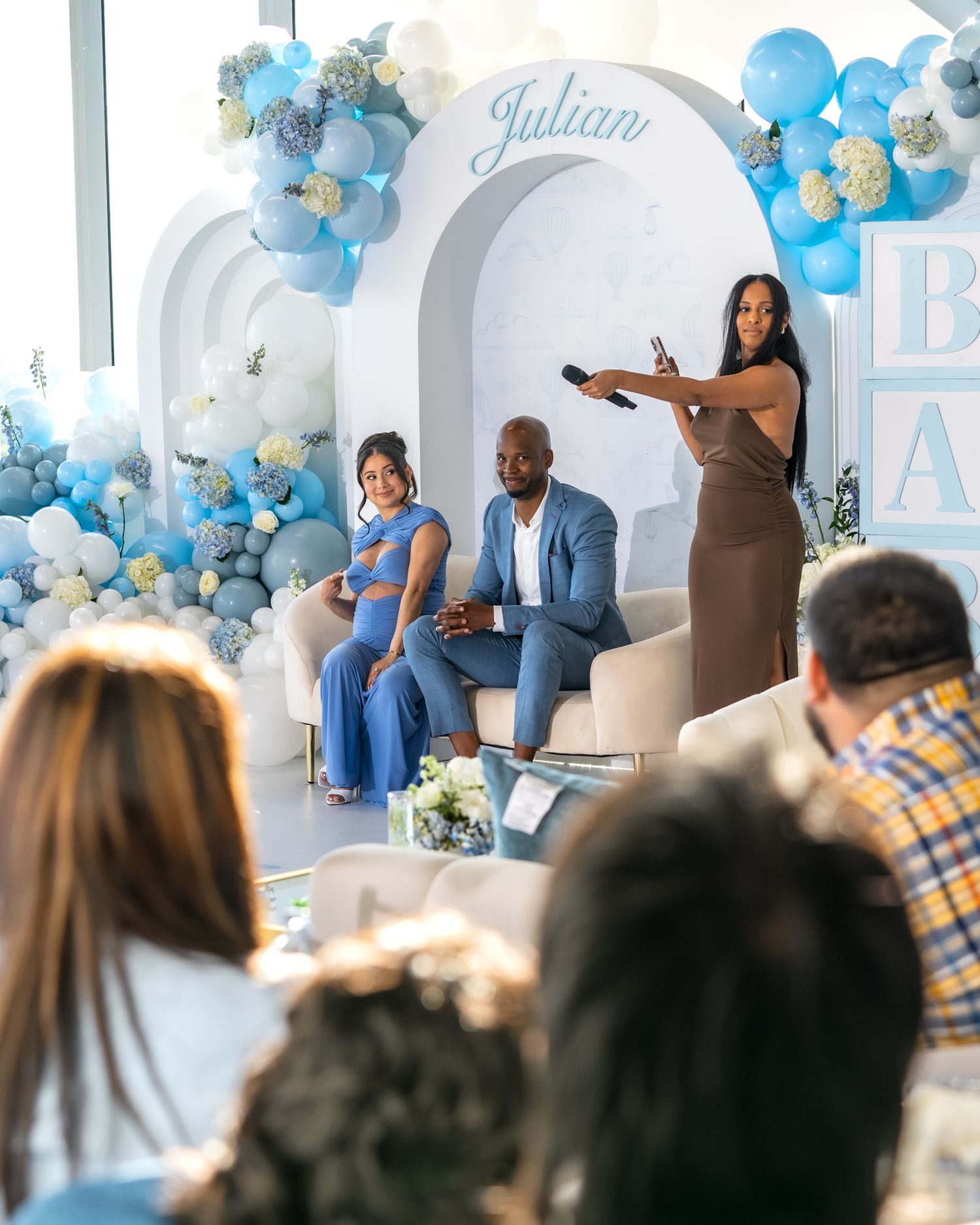 Baby shower celebration with guests seated watching presenters in front of decorative arch.