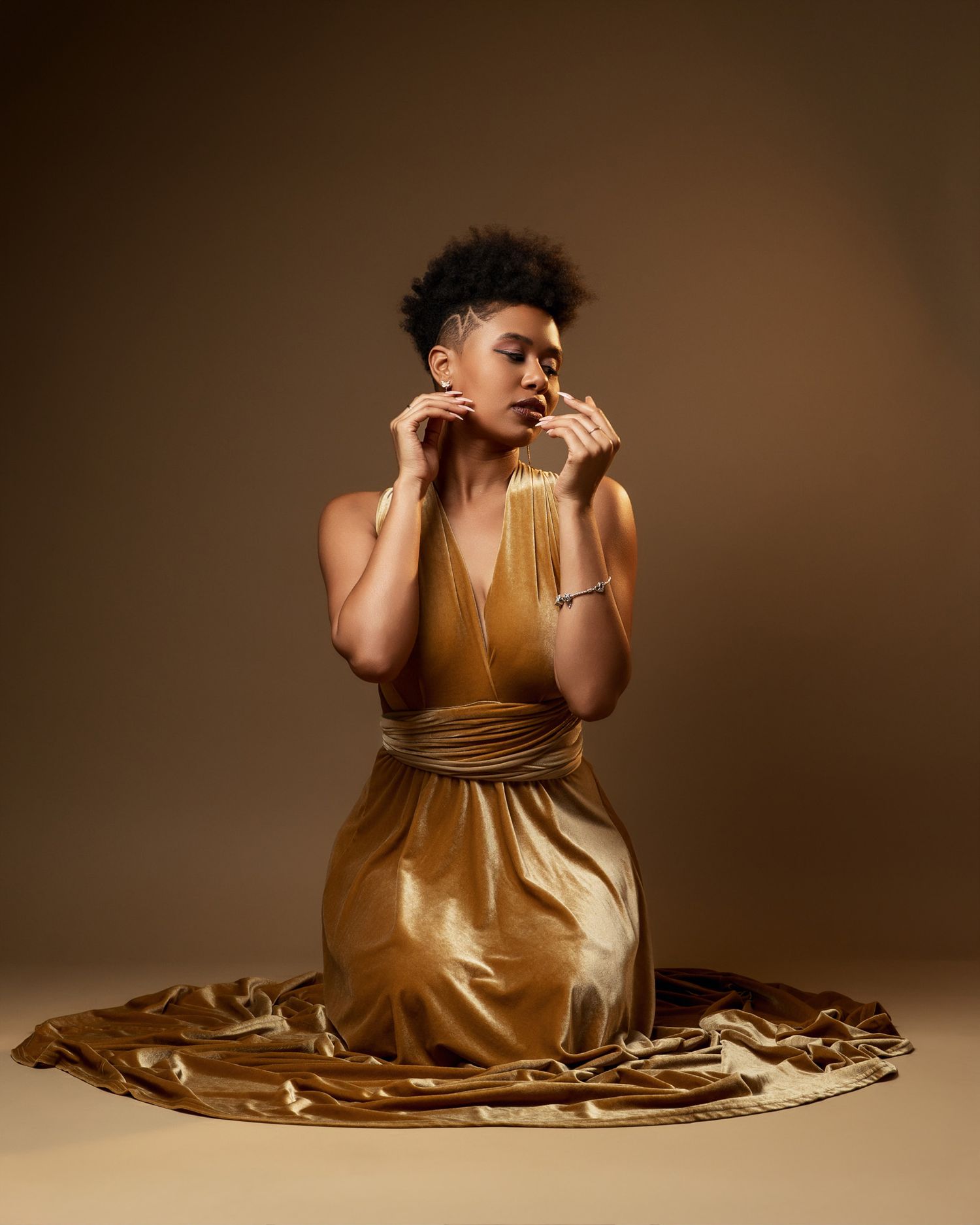 Elegant pose in metallic gold dress with hands touching face against brown backdrop.