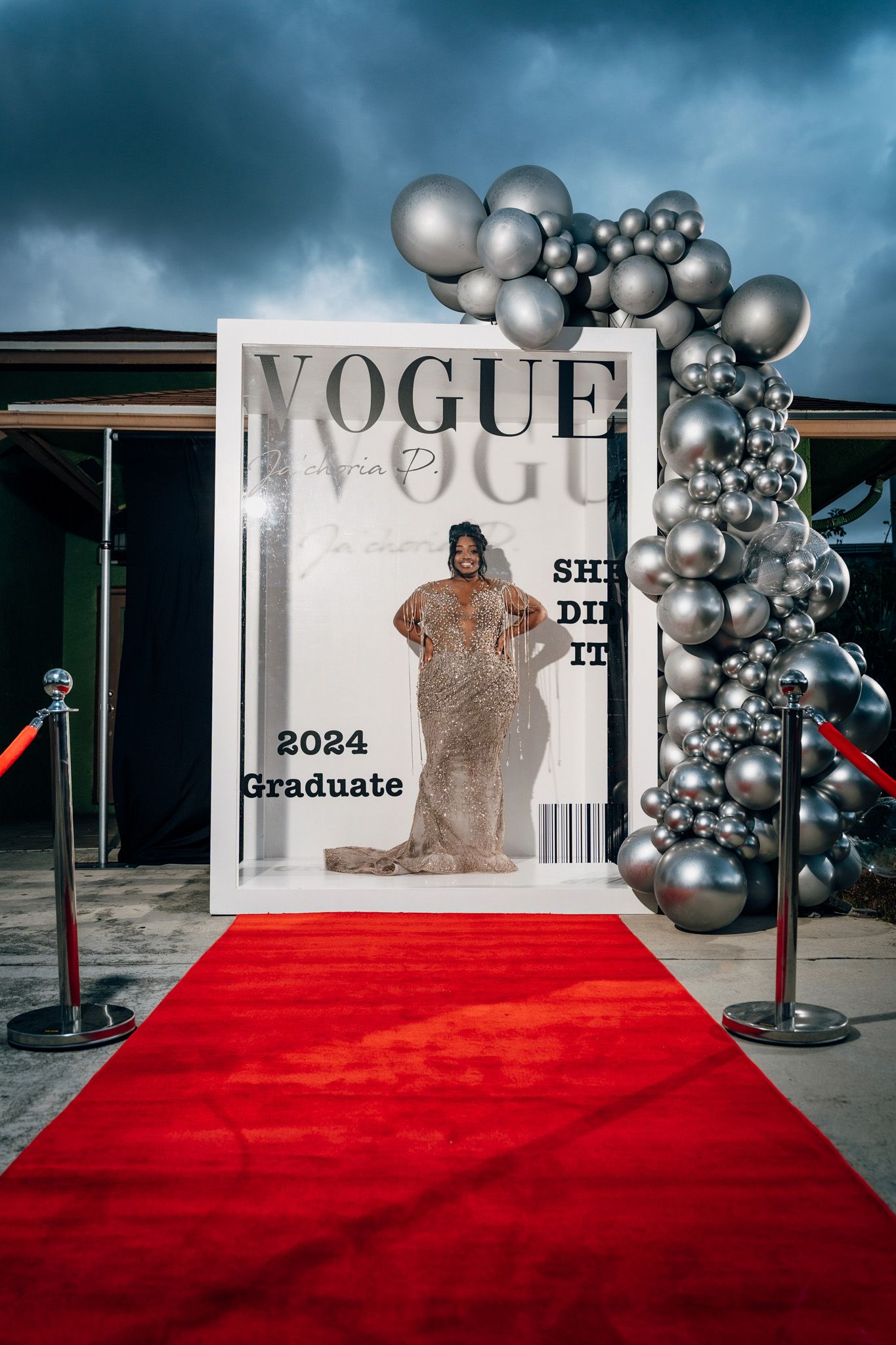 Wide shot of graduation photo setup with large Vogue frame, red carpet and silver balloon decor.