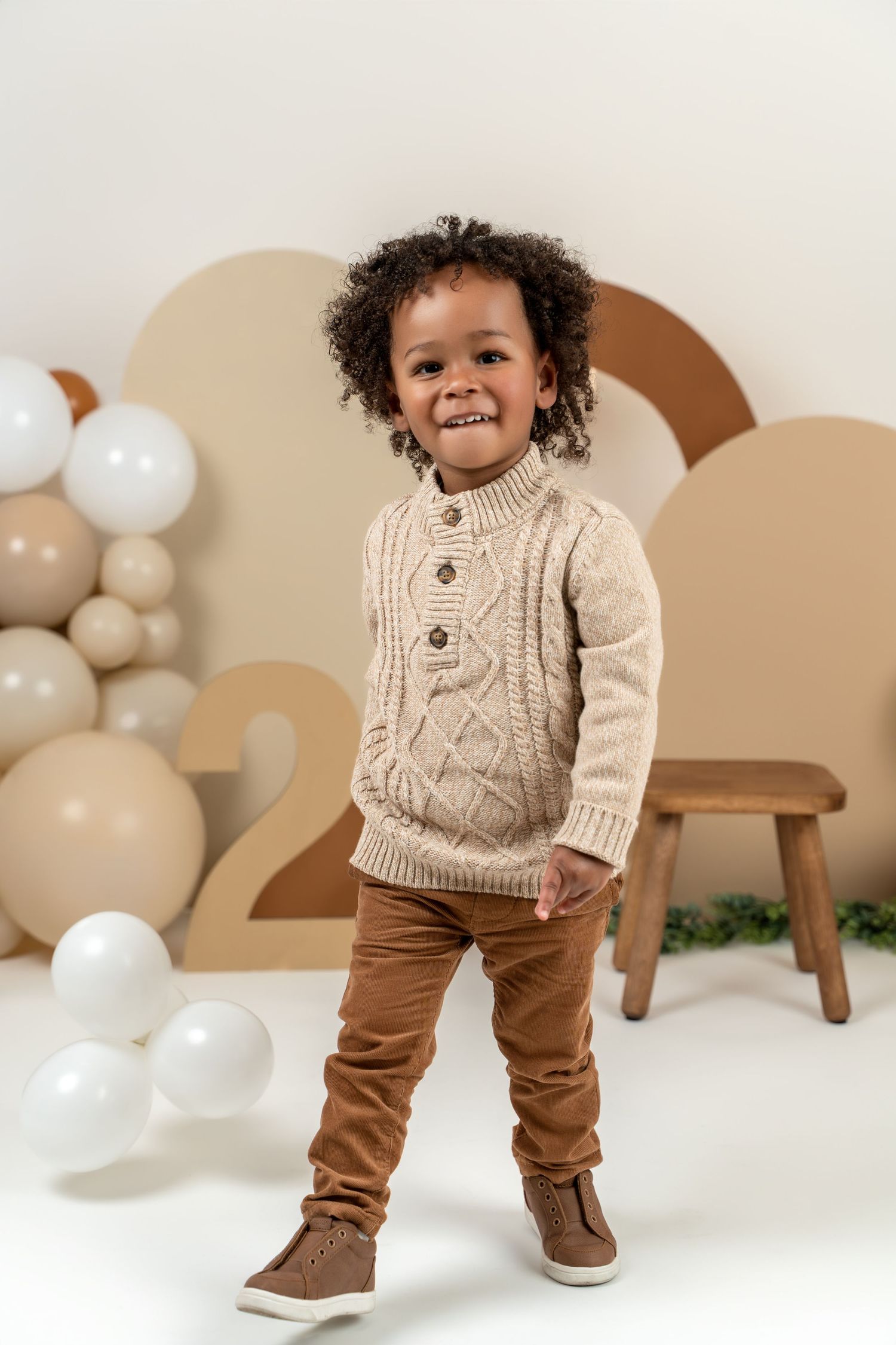 Small child wearing a cozy beige cable knit sweater and brown pants stands against a neutral studio backdrop with balloons.